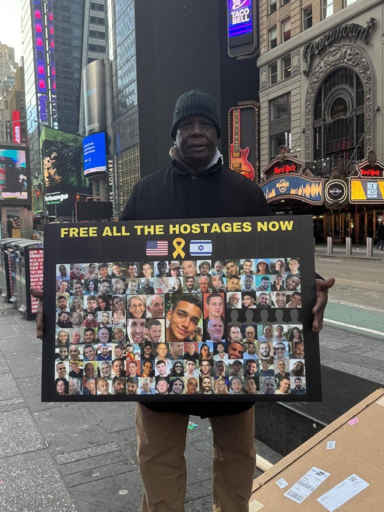 Simon Deng, a former child slave, standing in Times Square advocating for the release of hostages, holding a sign with images of people and wearing a solidarity dog tag.