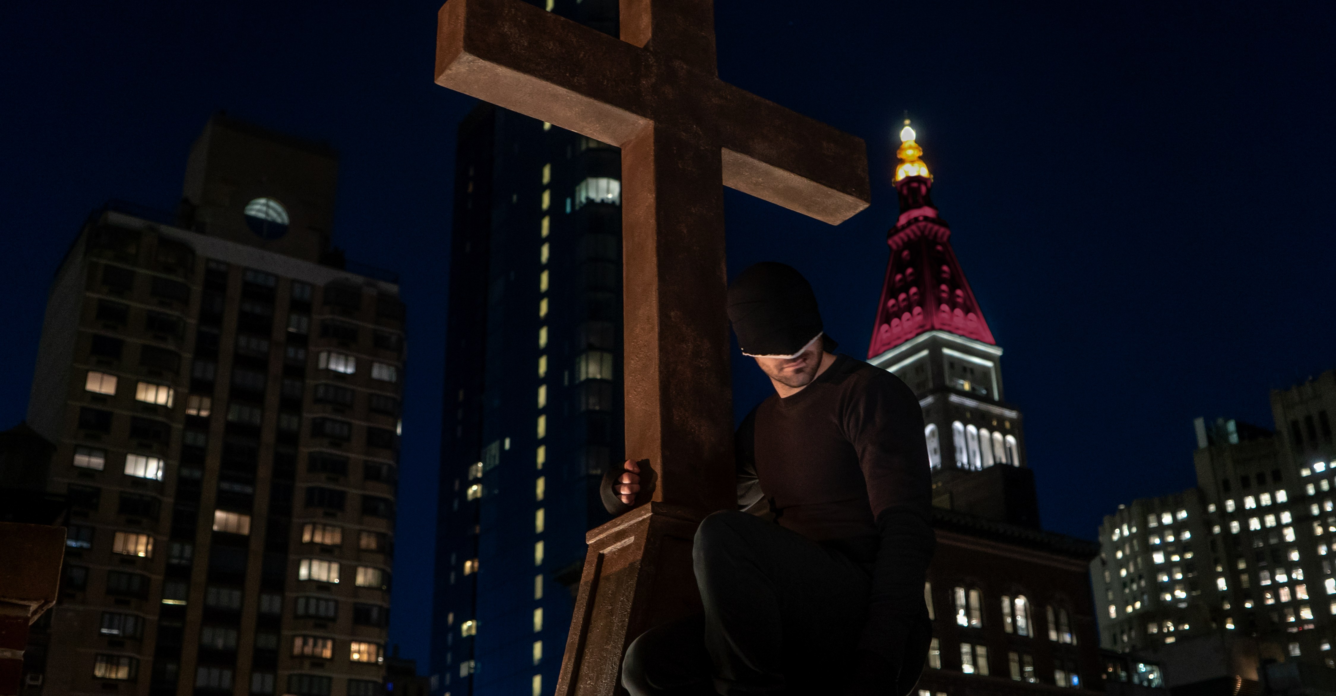 Daredevil in his black suit gripping a crucifix in front of the empire state building