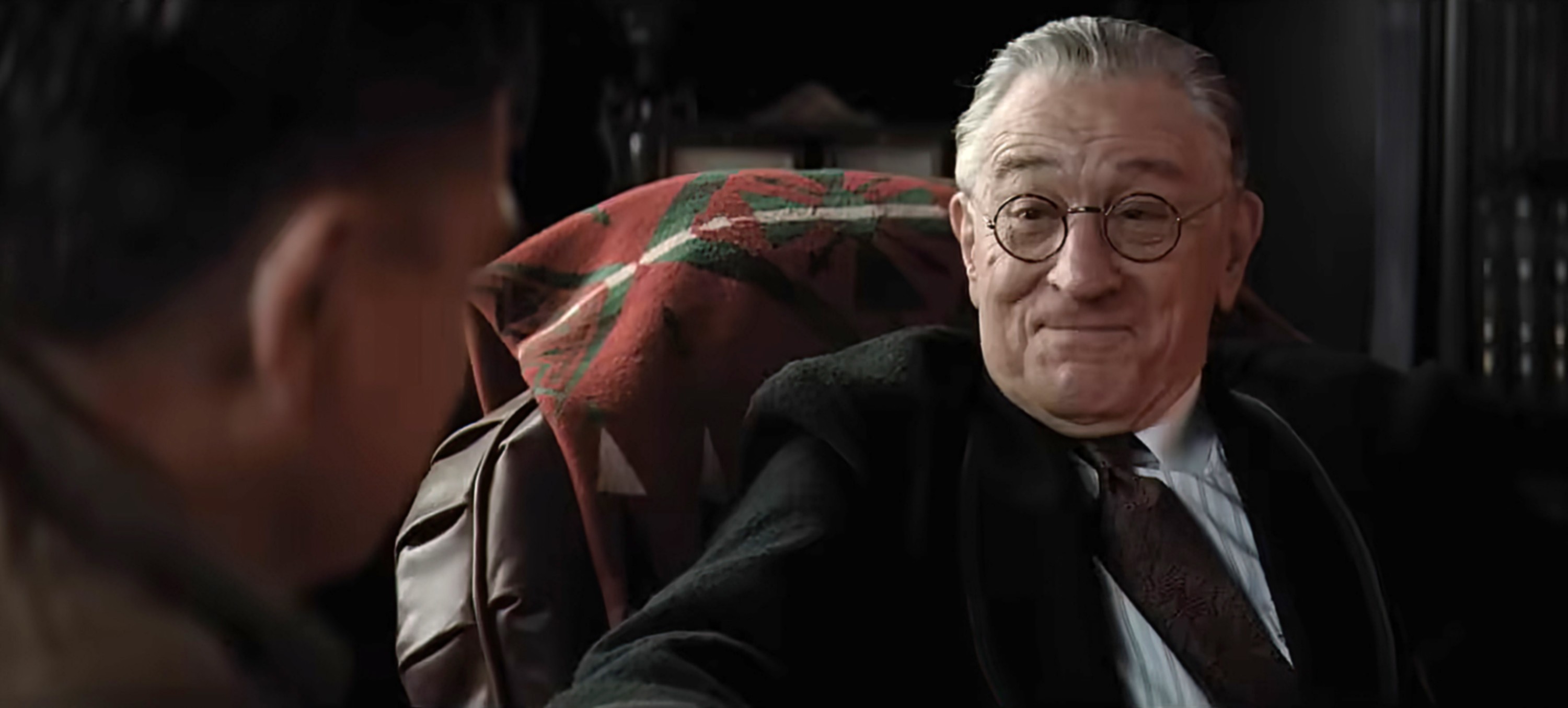 Robert De Niro sits in a chair wearing glasses. 