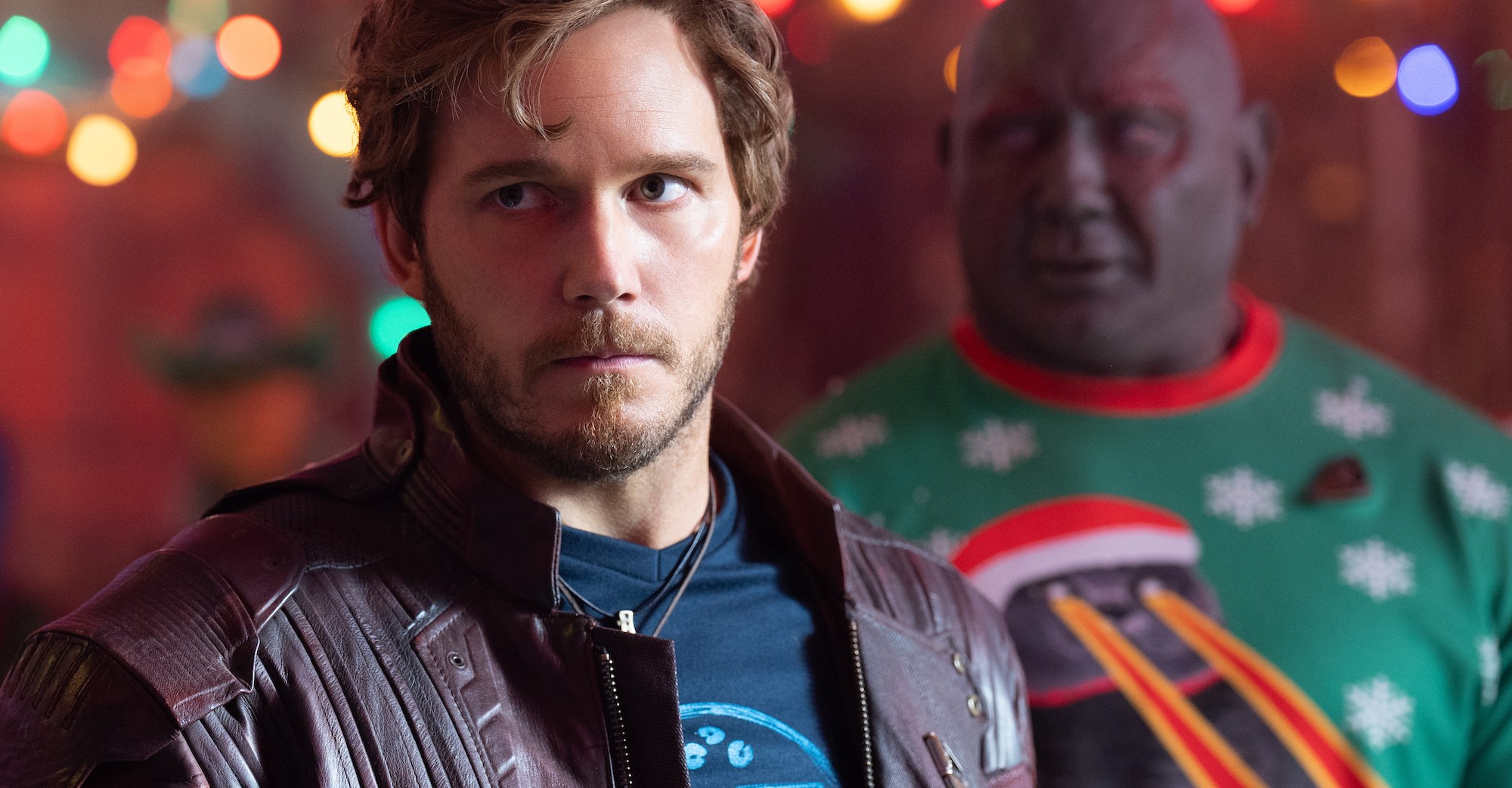 Human hero Peter Quill/Star-Lord looks frustrated in the foreground as his teammate Drax the Destroyer (Dave Bautista, in purple makeup, bright-red scarification designs, and a comically garish Christmas sweater depicting a creature with laser-eyes) smiles in the background in The Guardians of the Galaxy Holiday Special