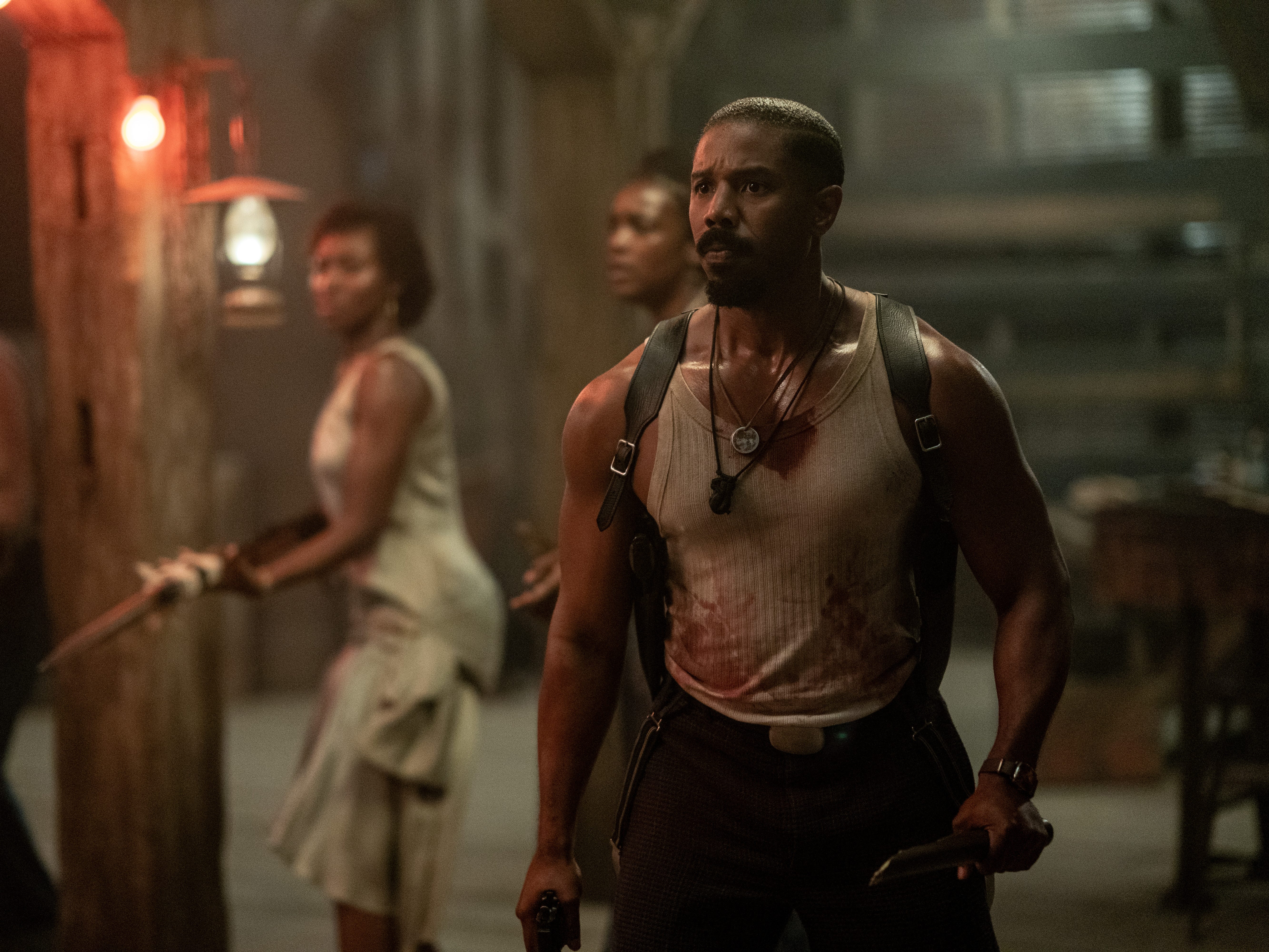 A still of "Sinners" showing Michael B. Jordan in a bloody vest, holding a broken wooden stick and gun with other people with weapons in the background.