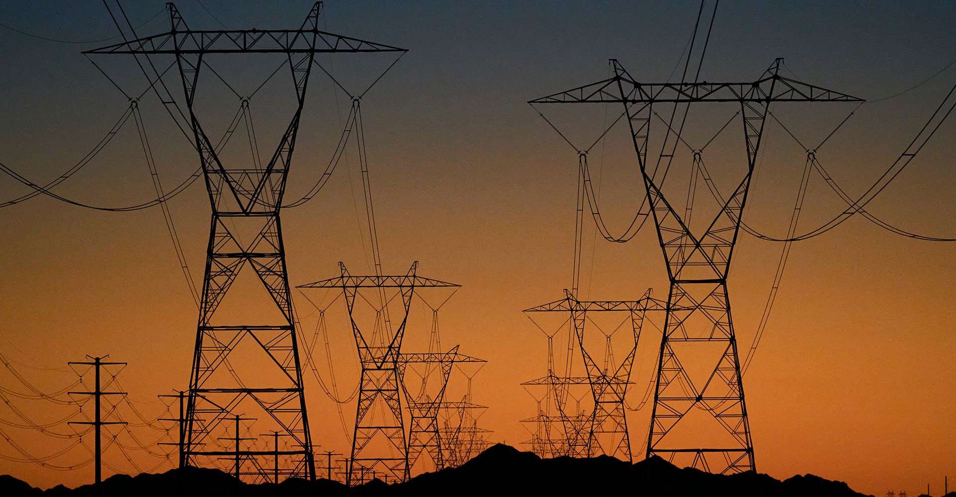 Massive power transmission towers seen beyond rocky terrain against a bright orange and blue sunset