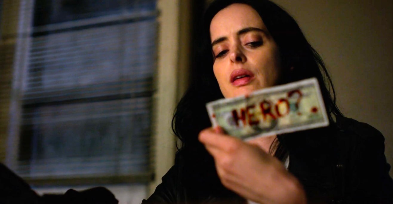 A screenshot of Jessica Jones from the Netflix series holding up a dollar bill onto which someone has scrawled the word “Hero?”