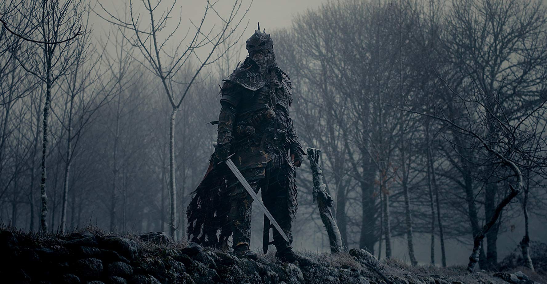 A warrior in black armor stands in front of a desolate winter forest.