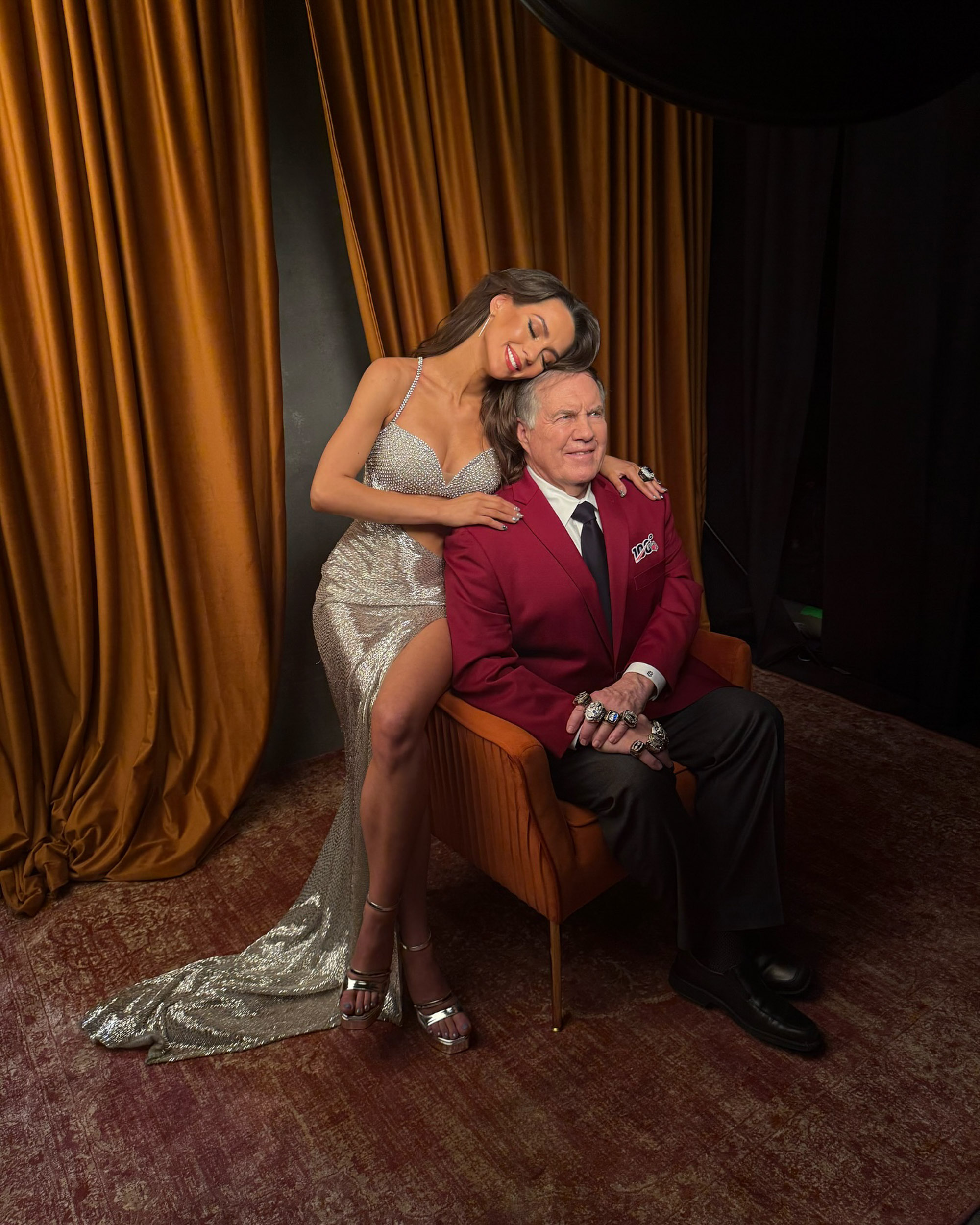 Jordon Hudson in a silver dress poses with Bill Belichick in a red suit coat. 