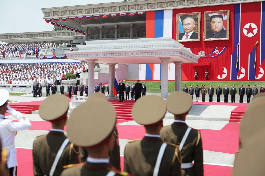 Kim Jong Un and Vladimir Putin standing under a stone gazebo with their portraits displayed in massive frames behind them. They are surrounded by people in military dress and others in suits.