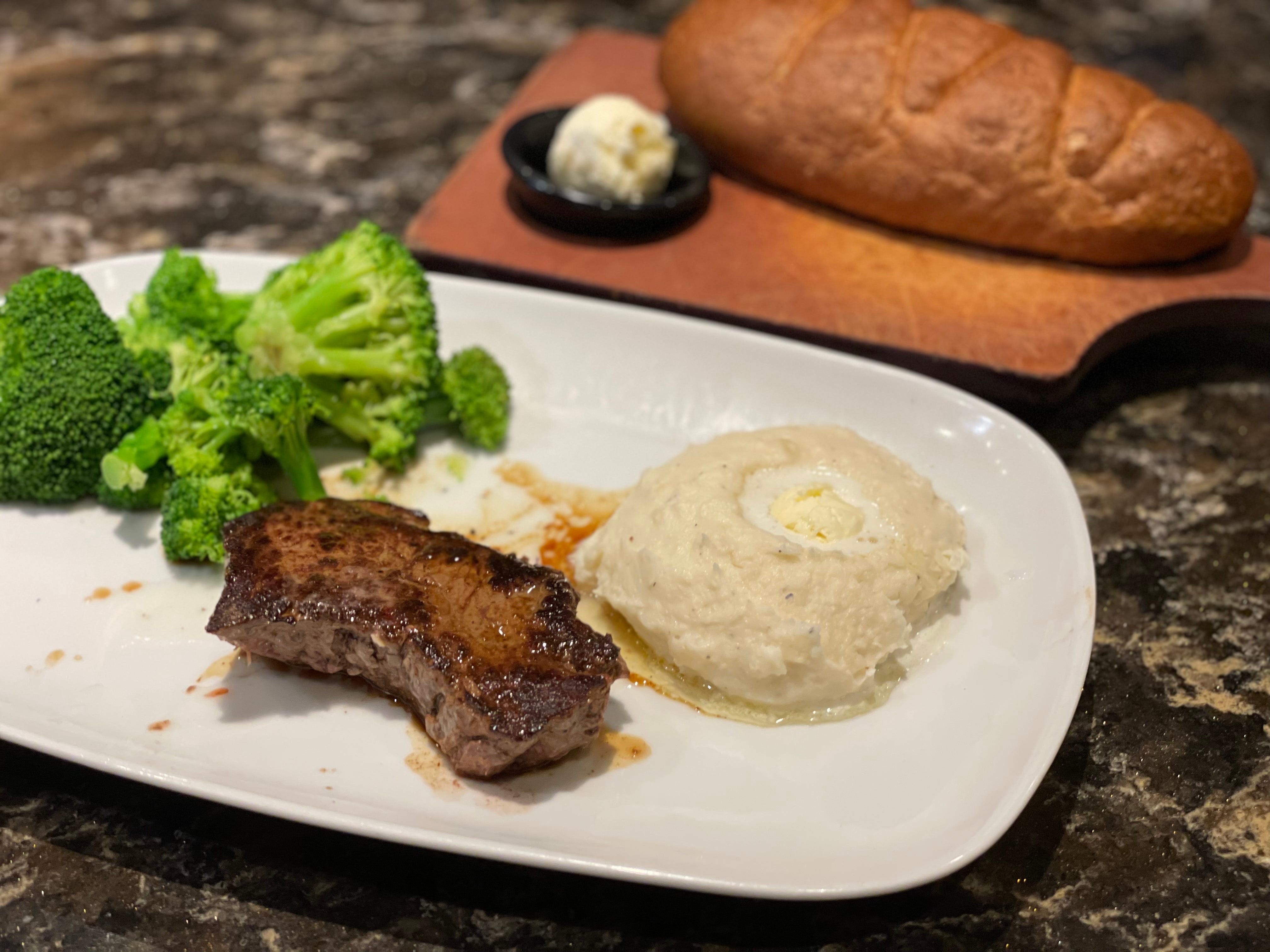 Steak, broccoli, mashed potatoes on plate with bread behind it at LongHorn Steakhouse