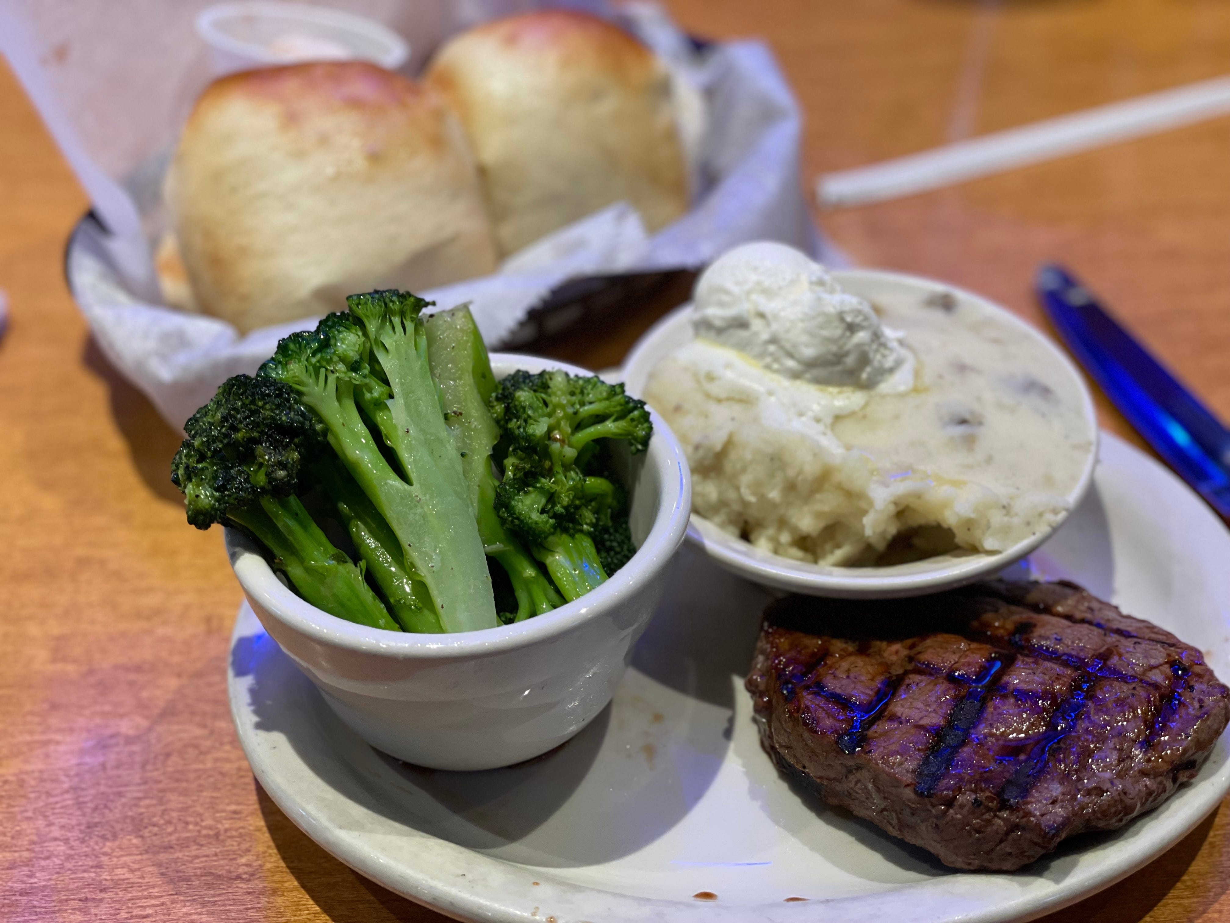 Steak on plate with bowls of mashed potatoes and broccoli and rolls behind them at Texas Roadhouse