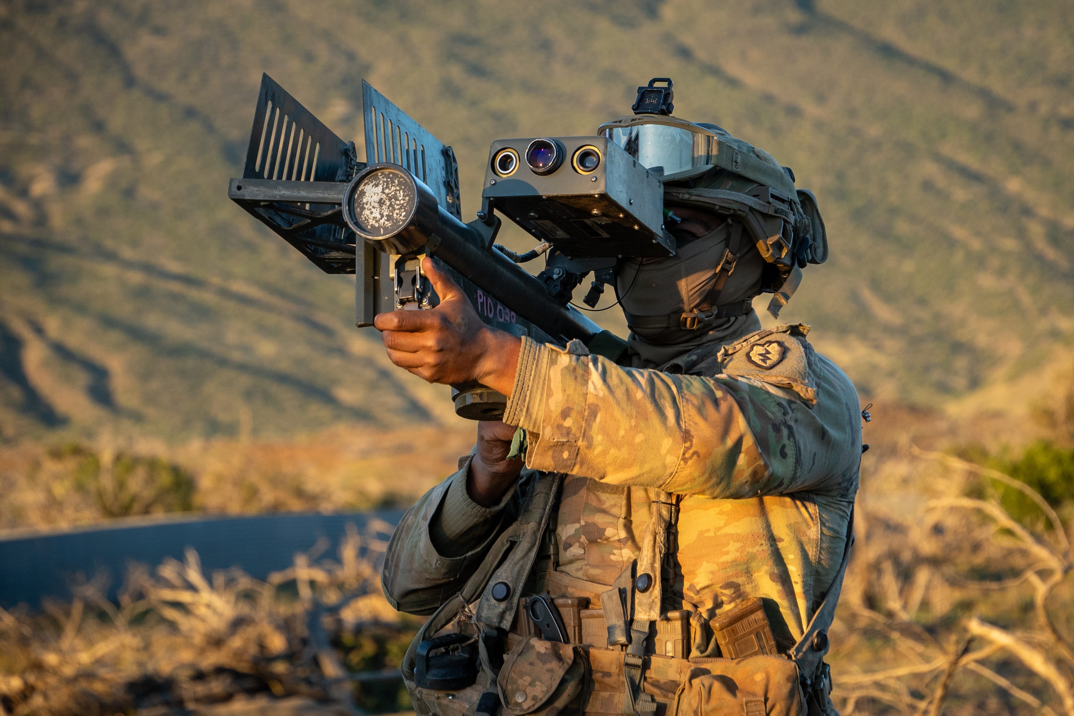 A US Army soldier wearing camouflage aims a weapon with a scope attached towards the sky, with a brown and green grassy background.