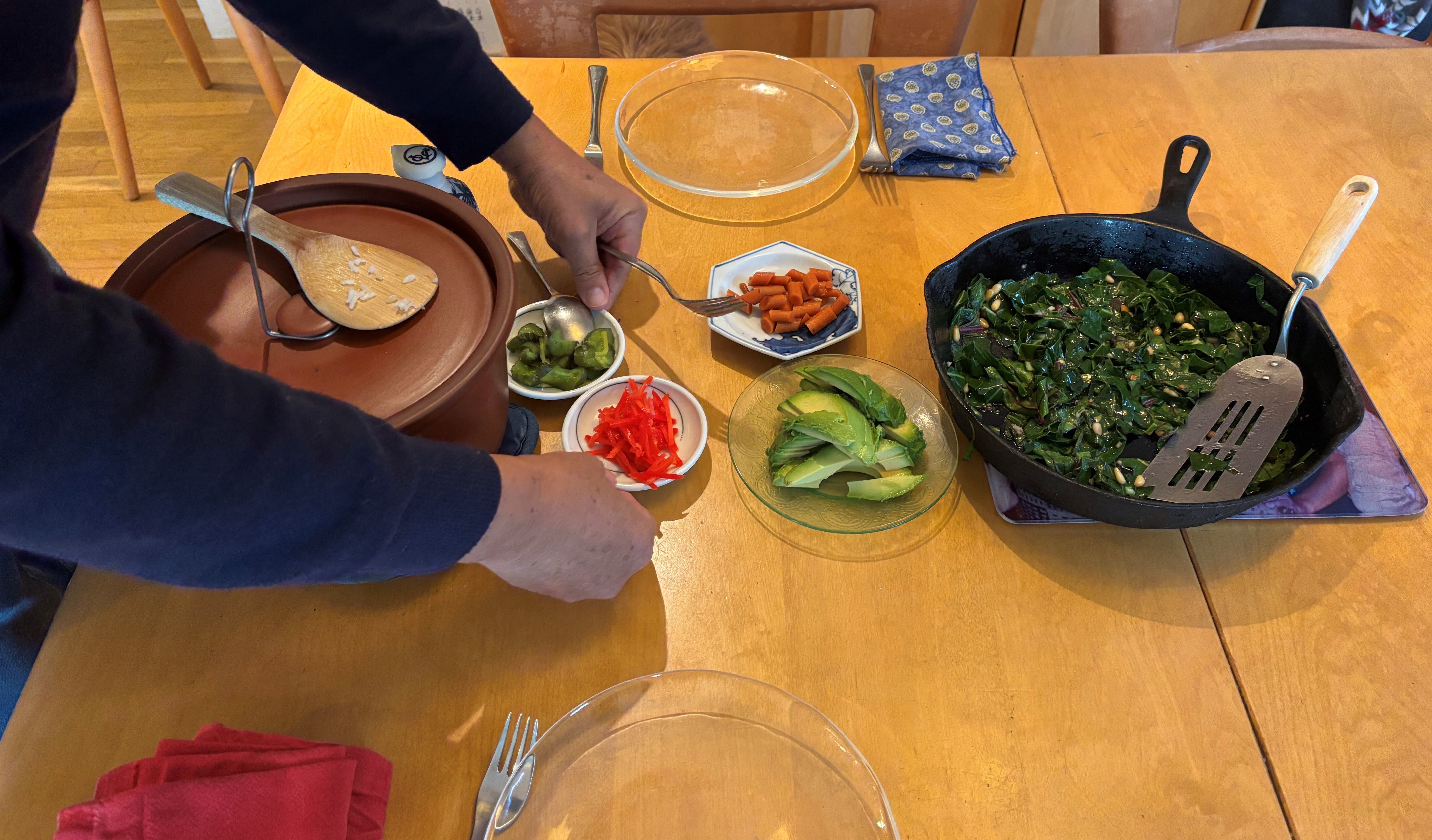 someone setting two small dishes of vegetables on a table set with two glass plates a large brown clay dish and a cast iron full of cooked leafy greens