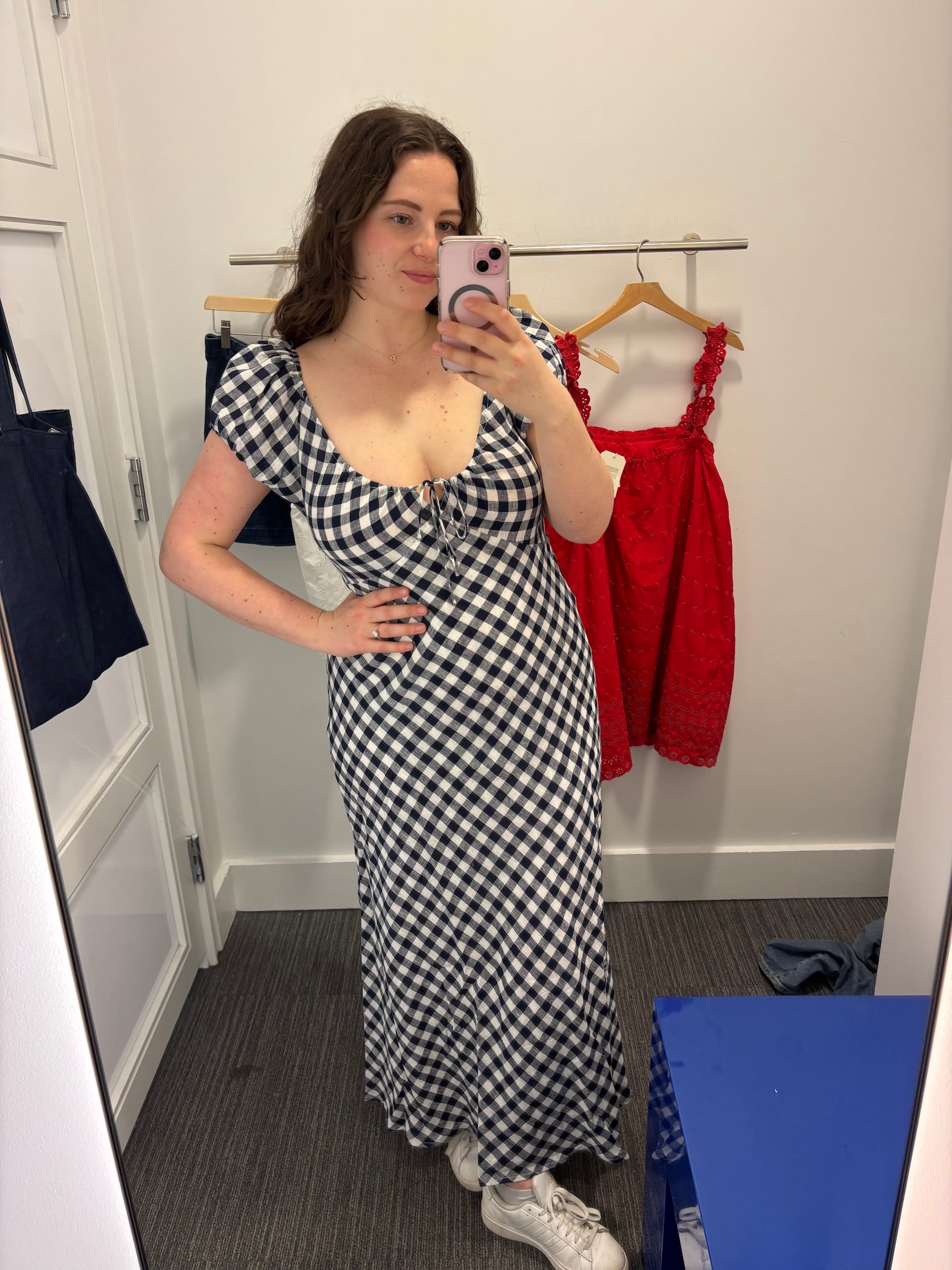 A woman in a gingham dress takes a mirror selfie in a dressing room.