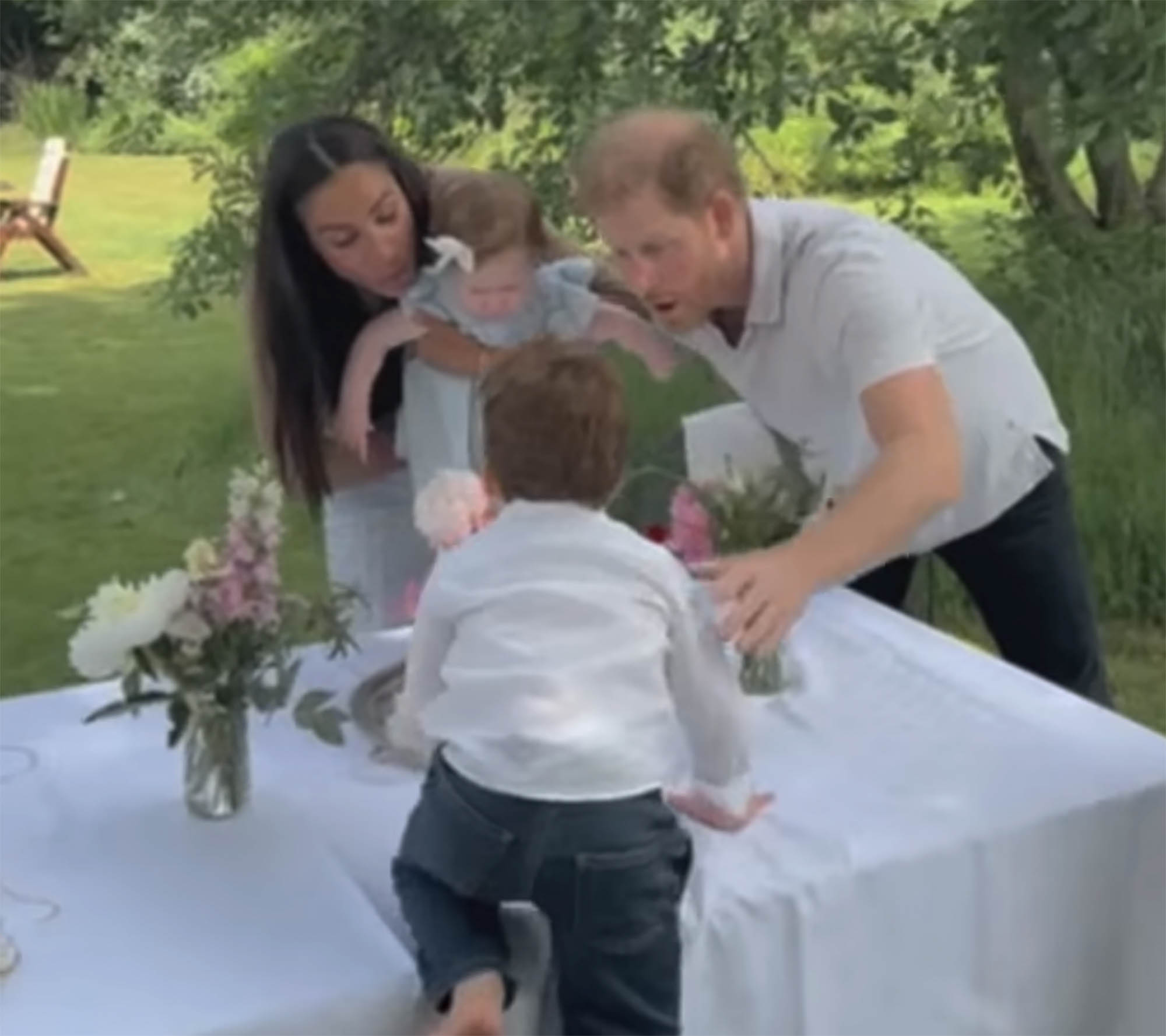 Prince Harry, Meghan Markle, and their two children gather around a table outdoors. 