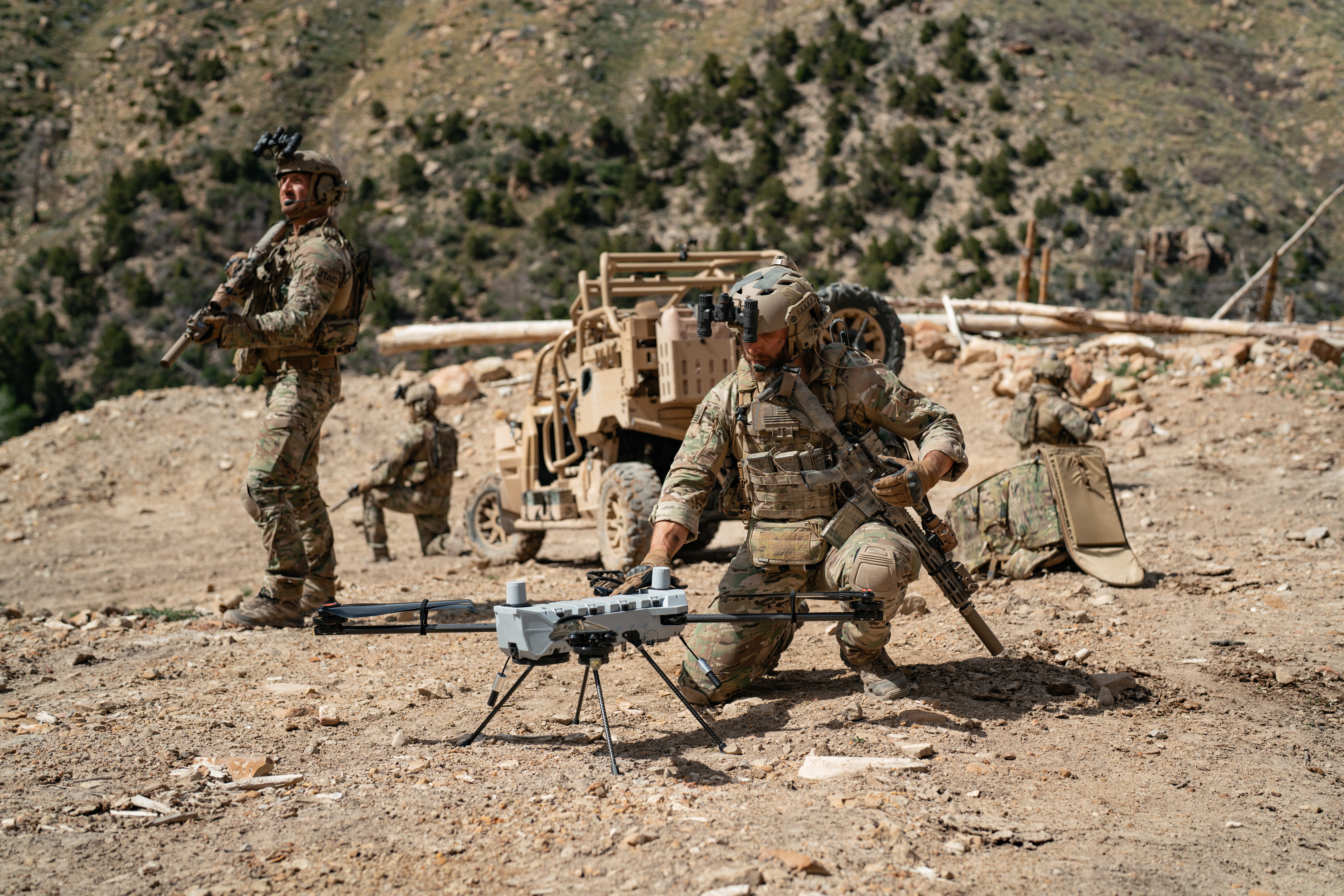 A group of US soldiers wearing camouflage stand around in dirt with a PDW C100 white drone on the ground.