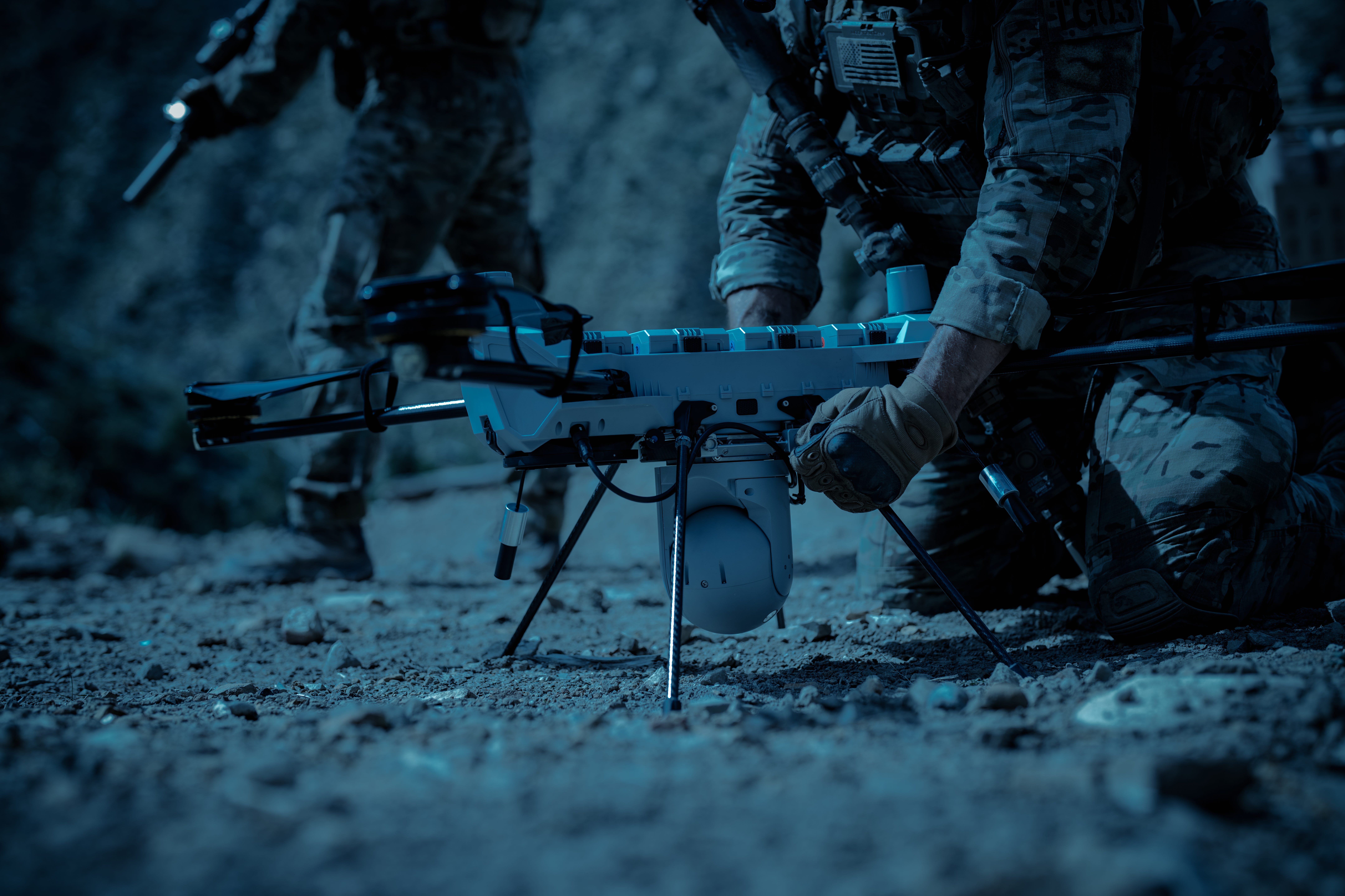 Two US soldiers wearing camouflage hold a PDW C100 drone on the dirt ground at night in darkness.