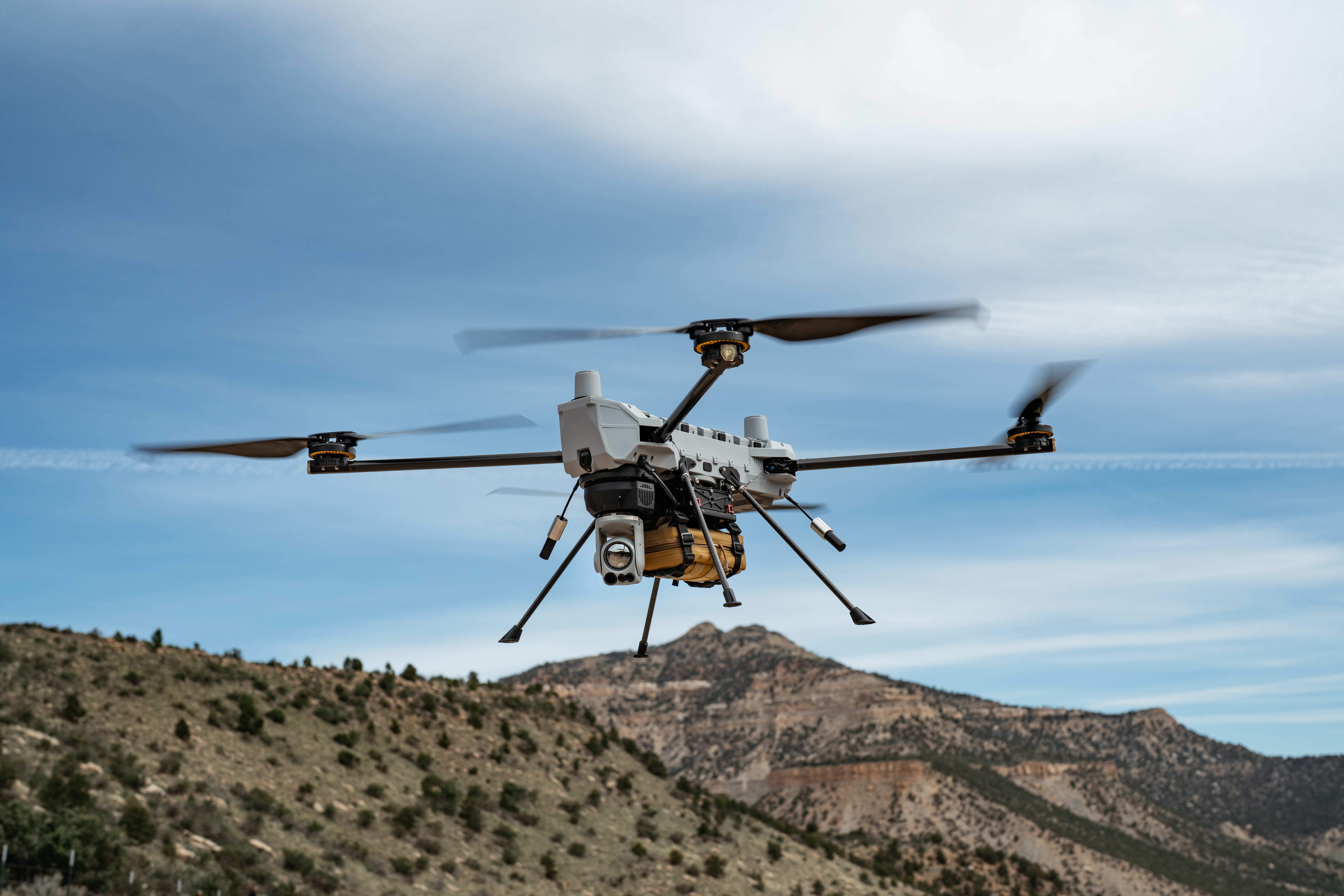 A C100 drone flies against a cloudy blue sky and greenish brownish mountains.