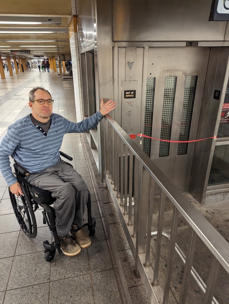 Sasha Blair-Goldensohn in a wheelchair in front of an out of service elevator, frustratingly pointing at the situation