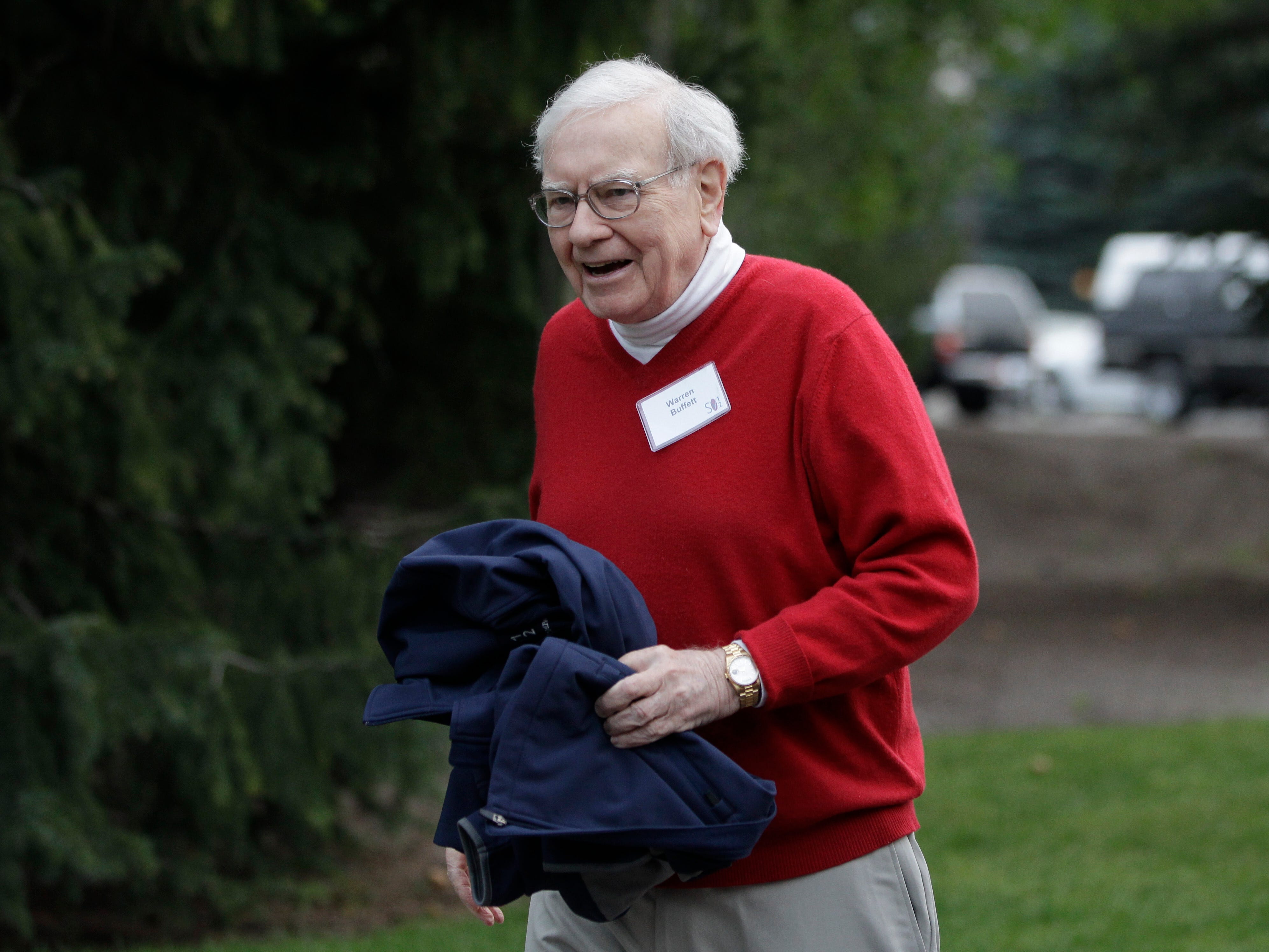Warren Buffett wearing a red shirt and holding a blue coat next to a hedge that it out of focus.
