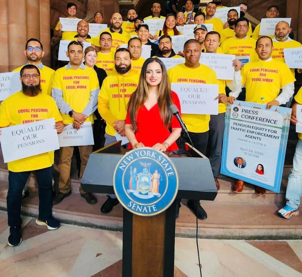 Jenifer Rajkumar at a state senate podium with a group behind her with yellow shirts reding equal pensions for equal work