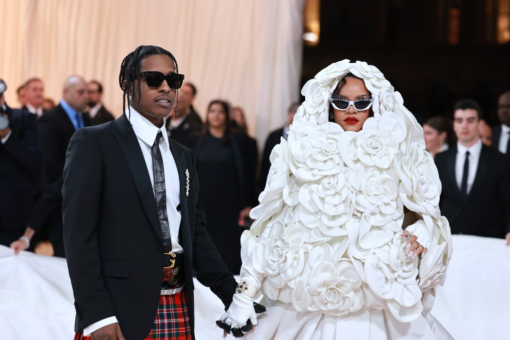 A$AP Rocky in a plaid skirt with a black blazer holding hands with Rihanna who is wearing a white headpiece dropped in flowers. 