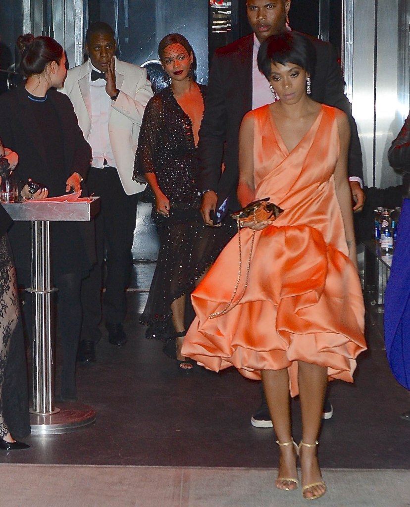 Solange Knowles in an orange dress walking out of the elevator with her sister Beyonce smiling in the background next to Jay Z. 