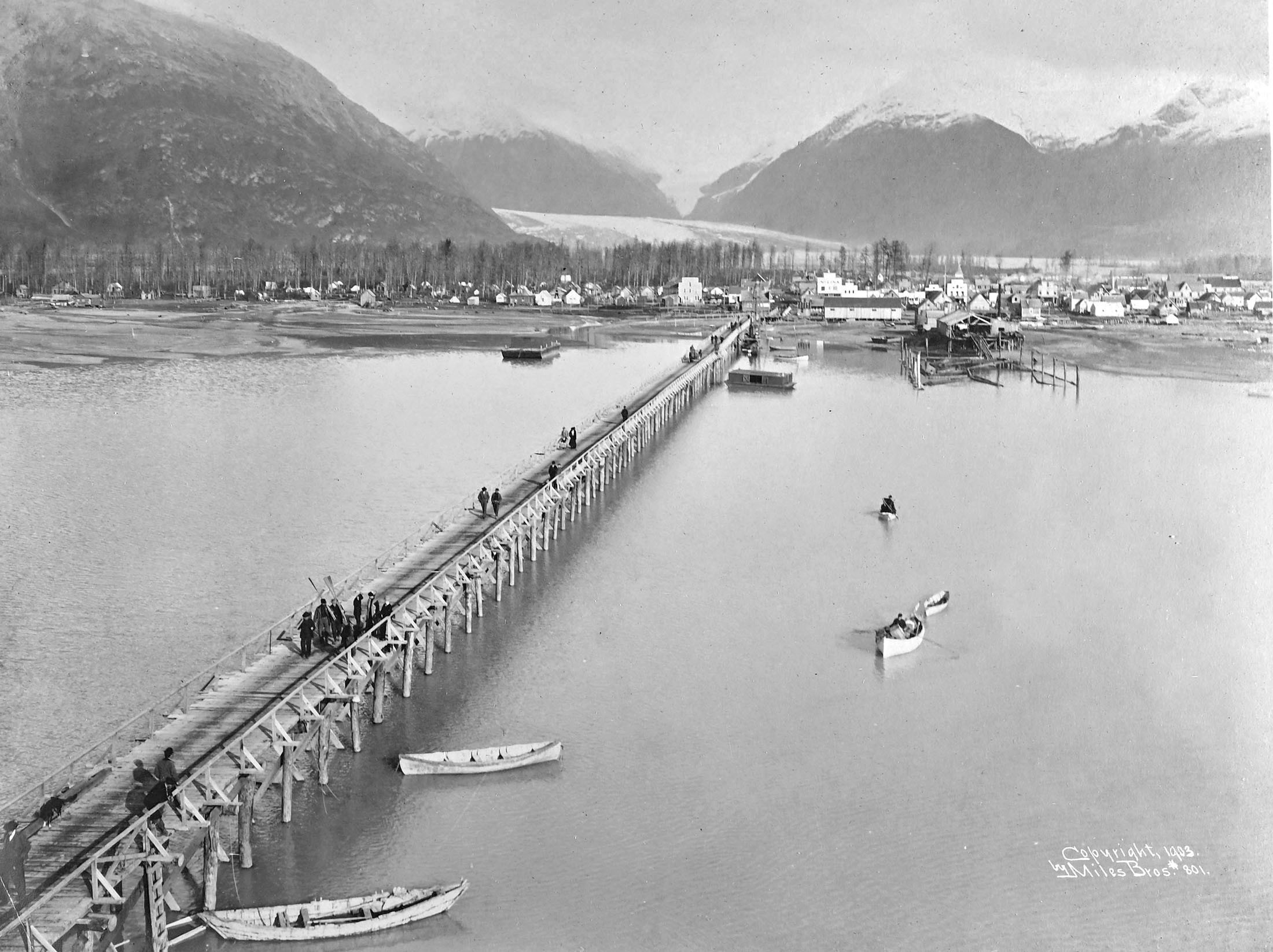A view from a tall ship anchored at a long pier, looking toward a small coastal town backdropped by mountains and a glacier.