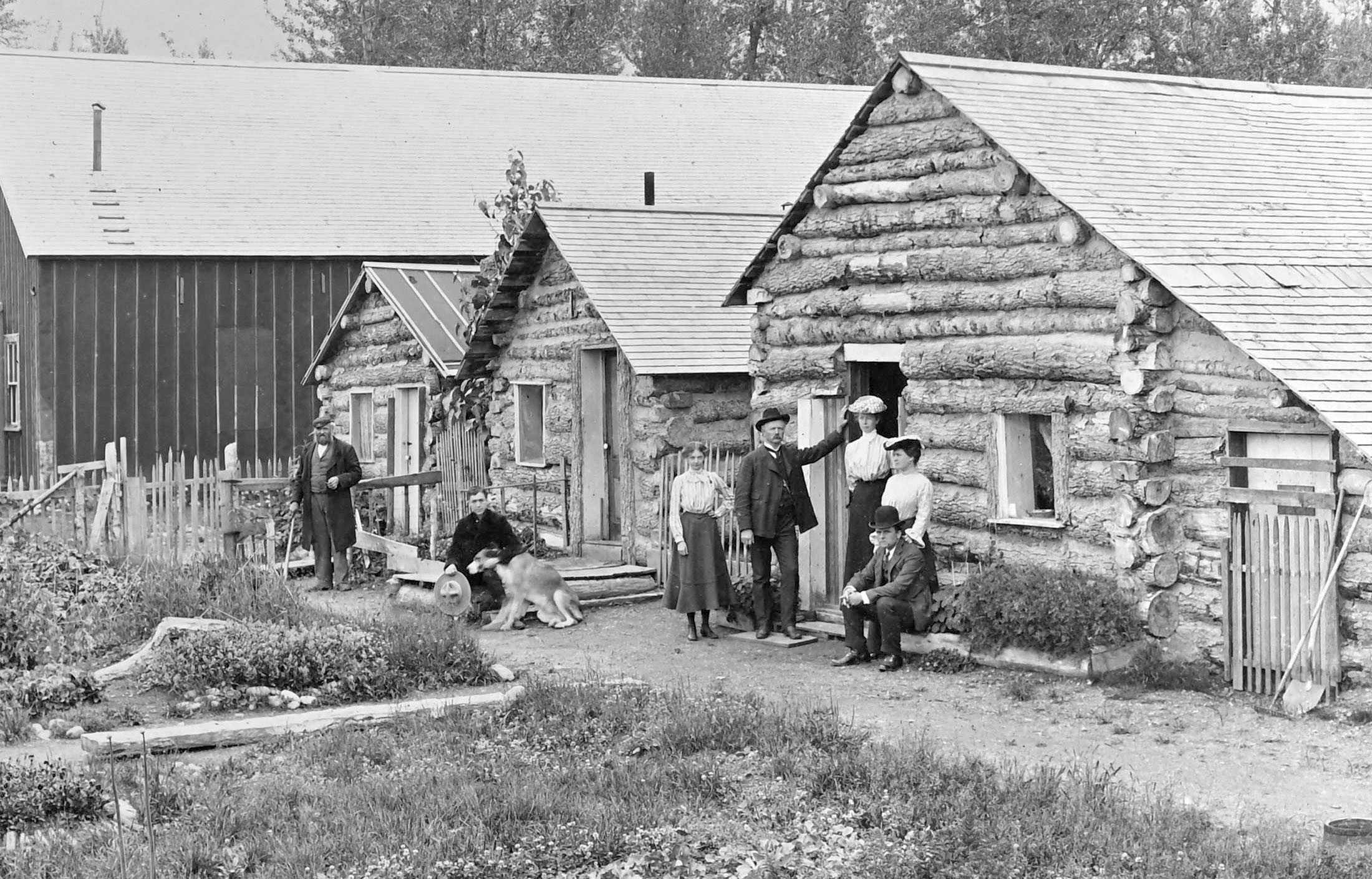 Seven men and women pose in front of a series of log cabins.