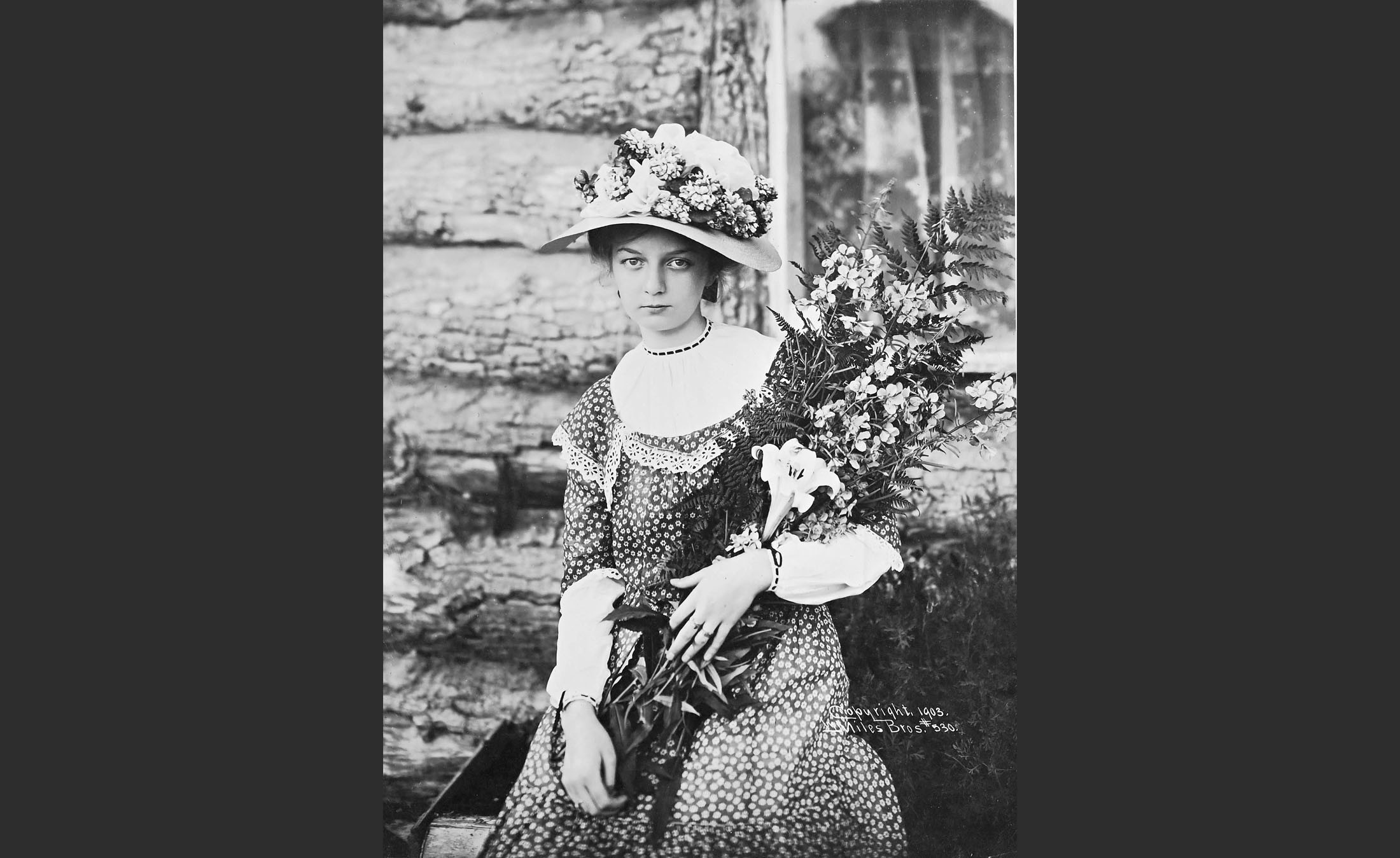 A young woman wearing a hat decorated with flowers, and holding a large bouquet.