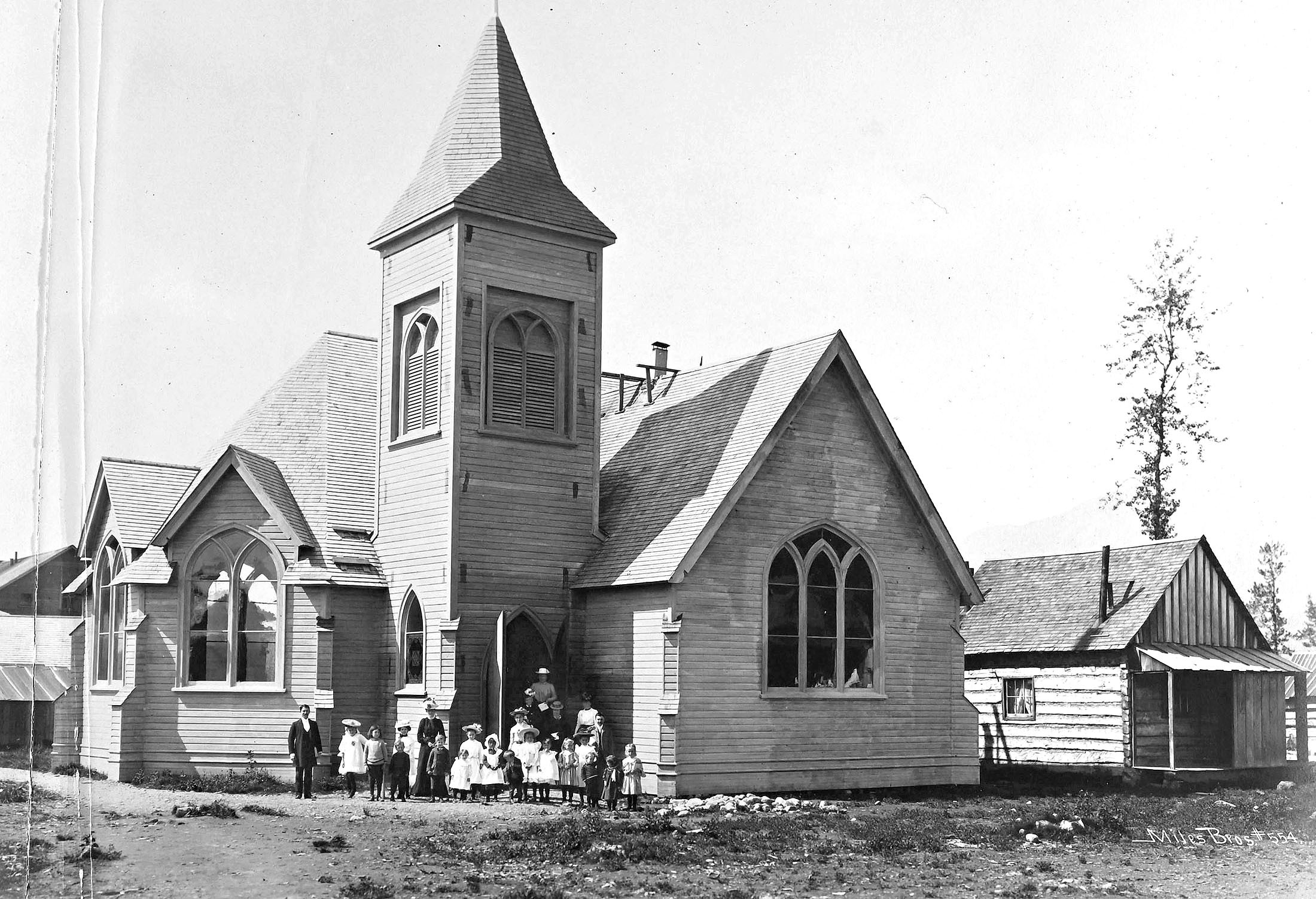 A group of well-dressed men, women, and children pose outside a wooden church.