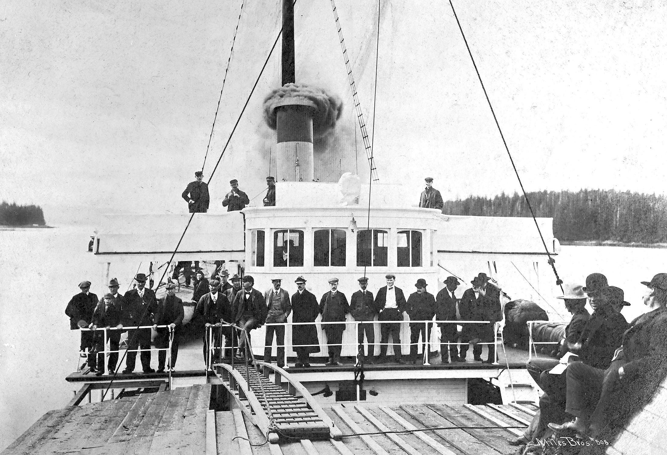 A couple of dozen people, mostly men, stand on the deck of a steamship sailing past forested islands.
