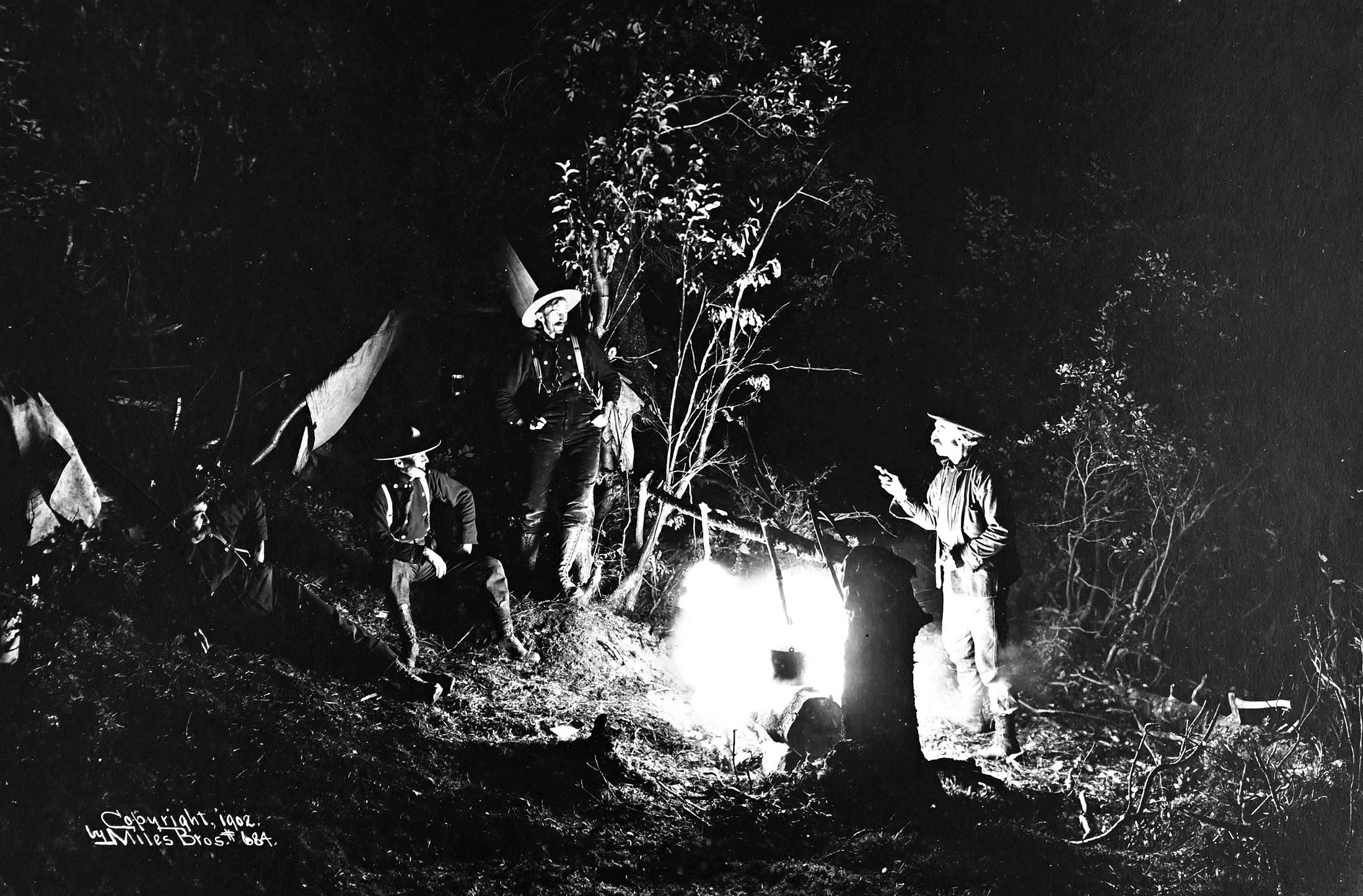 An older black-and-white photo of several men wearing broad-brimmed hats talking while gathered around a campfire at night