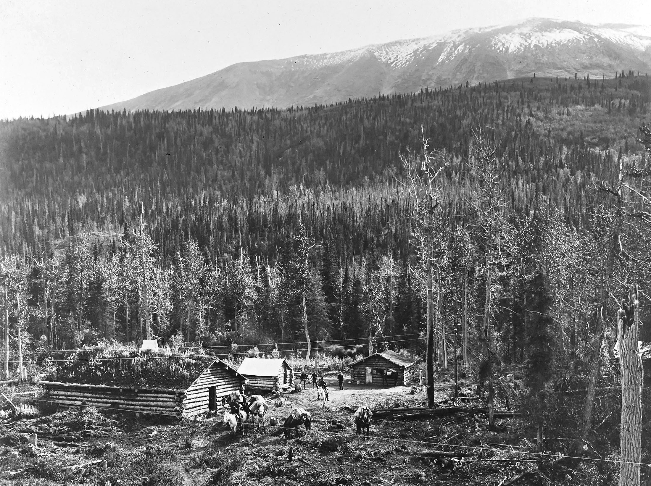 Several men and pack horses stand outside a log building, beneath a forested mountain slope.
