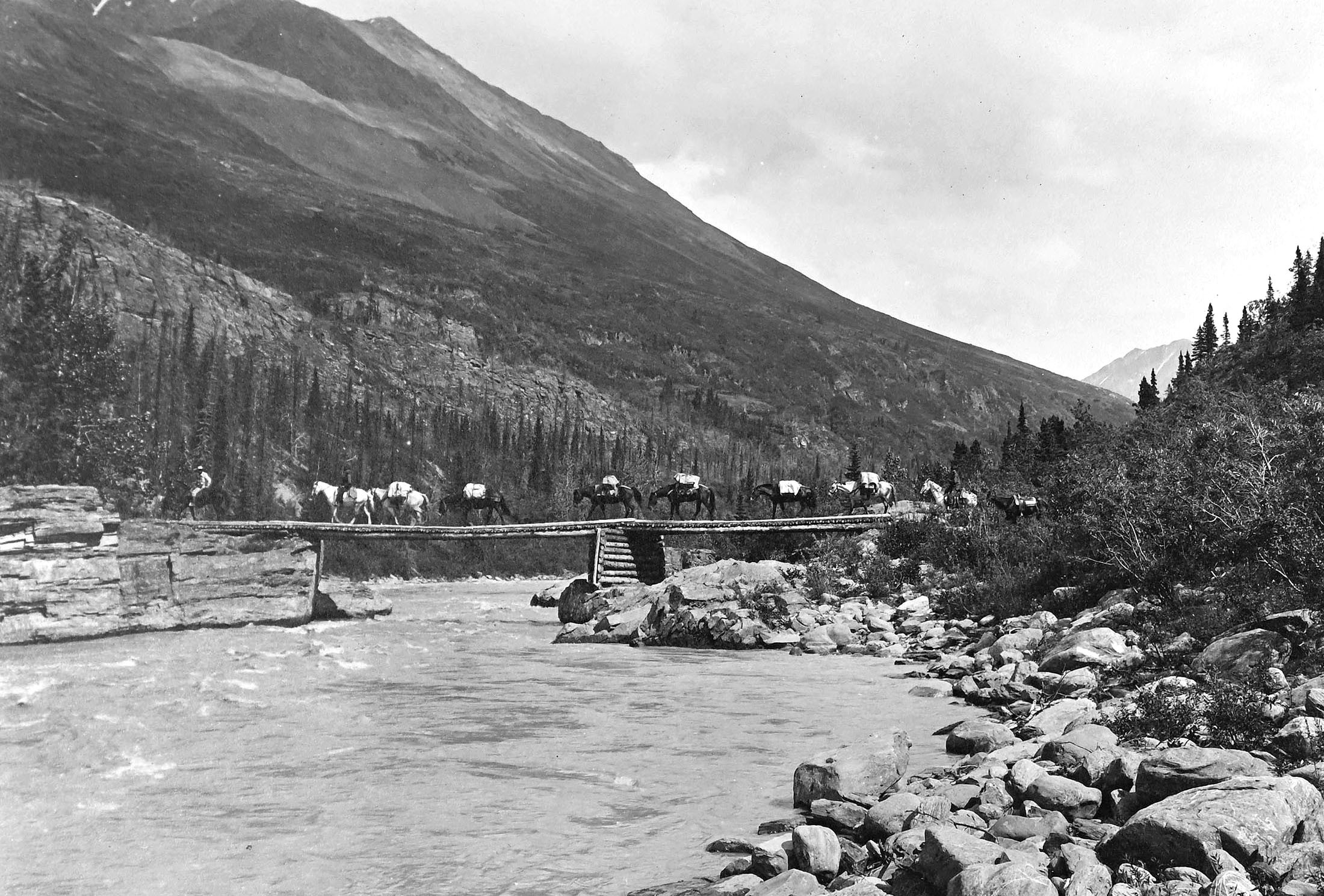 Three men ride horses, leading at least seven pack horses across a wooden bridge over a river.