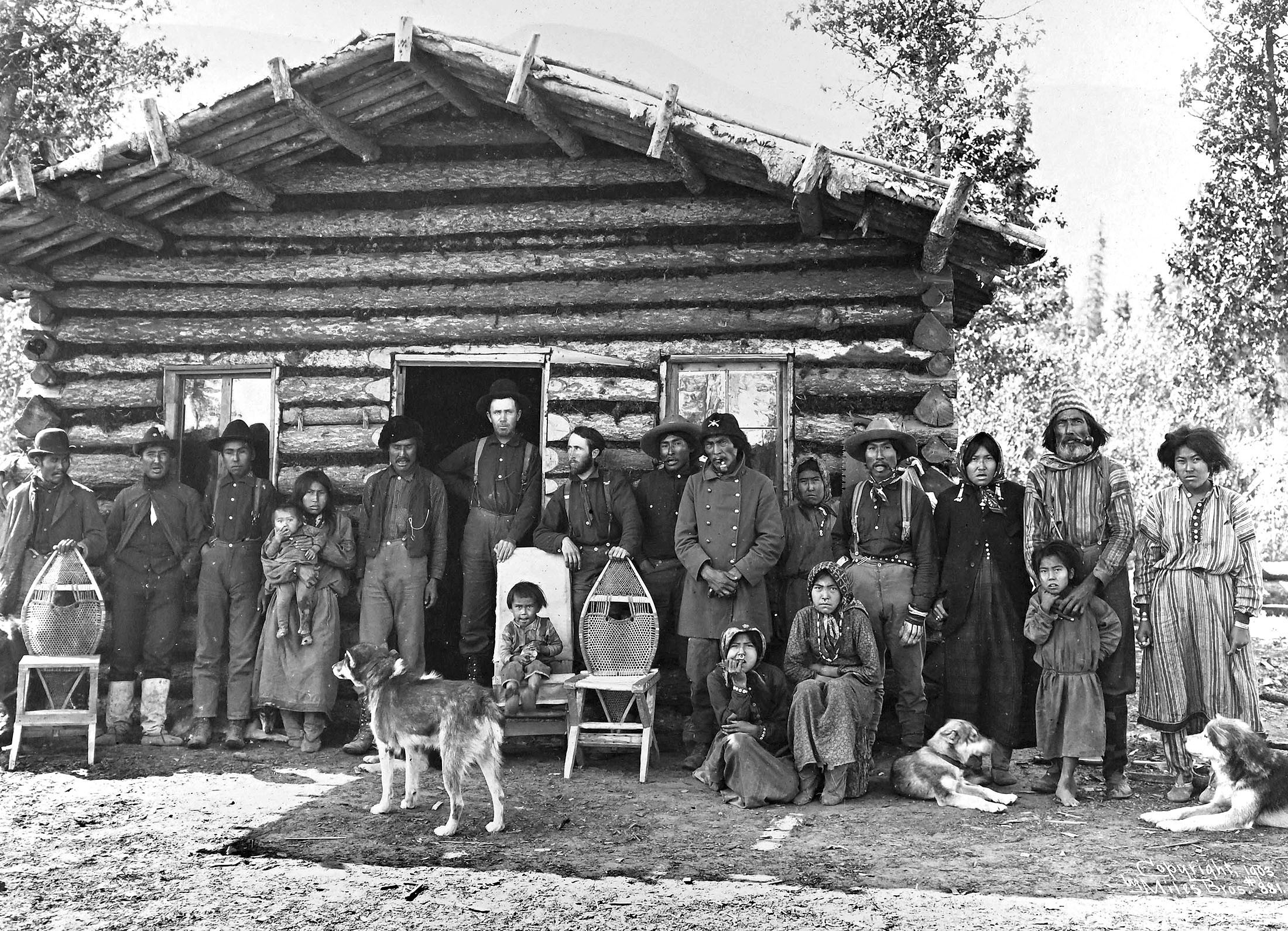 About twenty men, women, and children pose in a line, outside a log roadhouse.