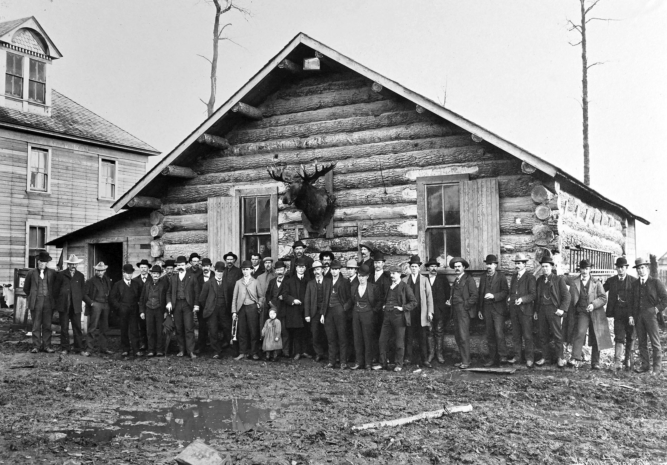 A large group of people pose side-by-side in front of a log building with a large moose head mounted on the outside.
