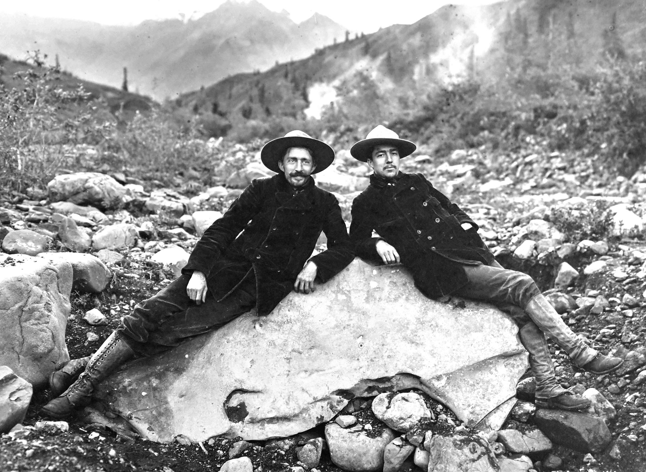 Two men pose while sitting on an enormous nugget of copper in a dry creek bed.