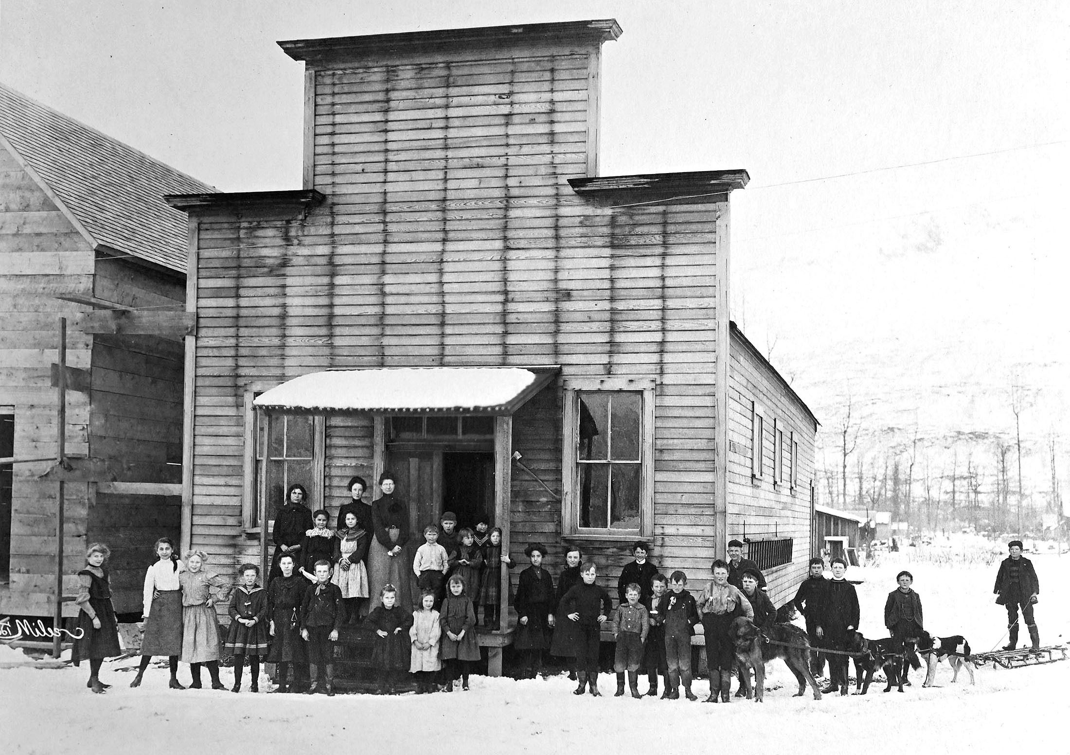 Schoolchildren, along with several sled-pulling dogs, pose in front of their school house.