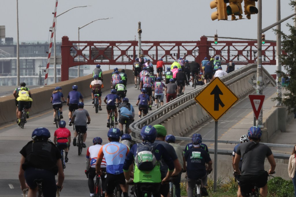 Thousands of cyclists participating in the 2025 Five Borough Bike Tour heading towards the Pulaski Bridge with the Queensboro Bridge in the background, escorted by the NYPD