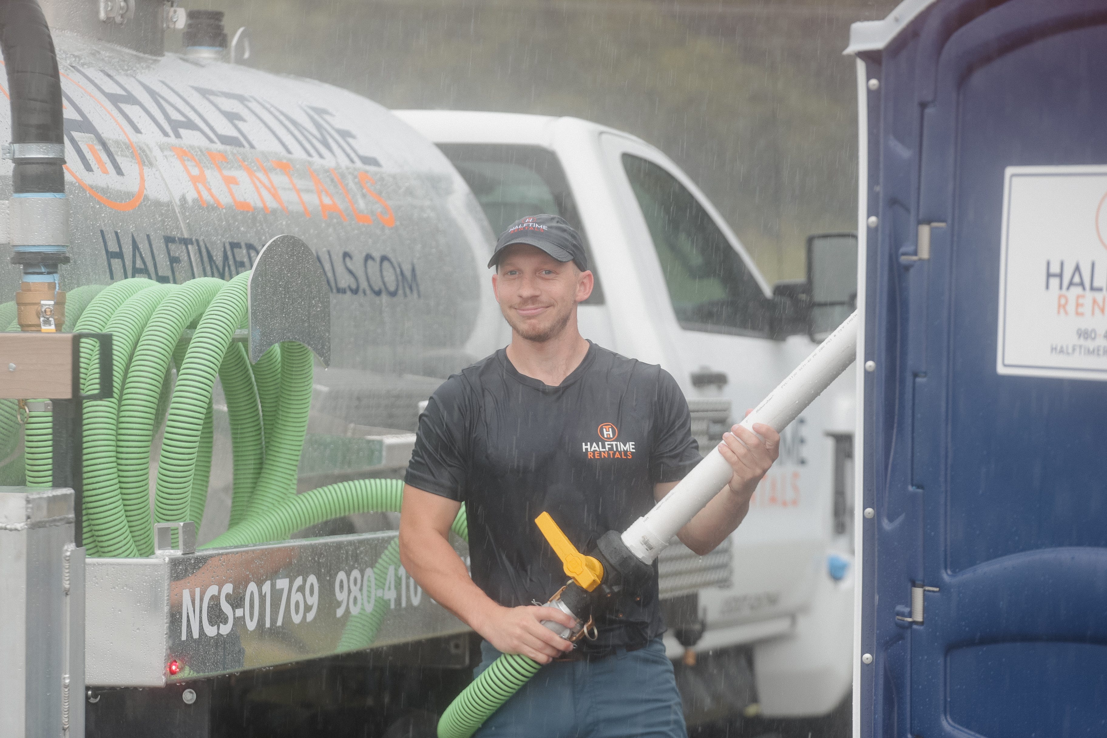 Man standing in the rain with a portable toilet truck and hose.