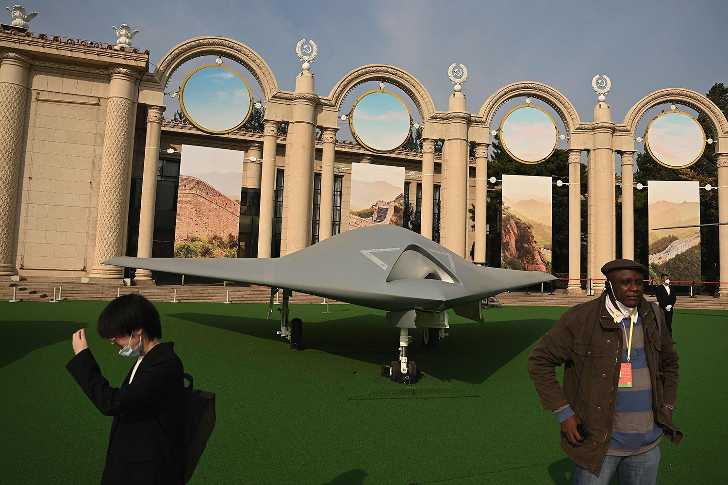 Two people wander past a large winged drone displayed at an open-air exhibit surrounded by a a wall made up of ornate columns and archways. A hazy sky is partly visible overhead.