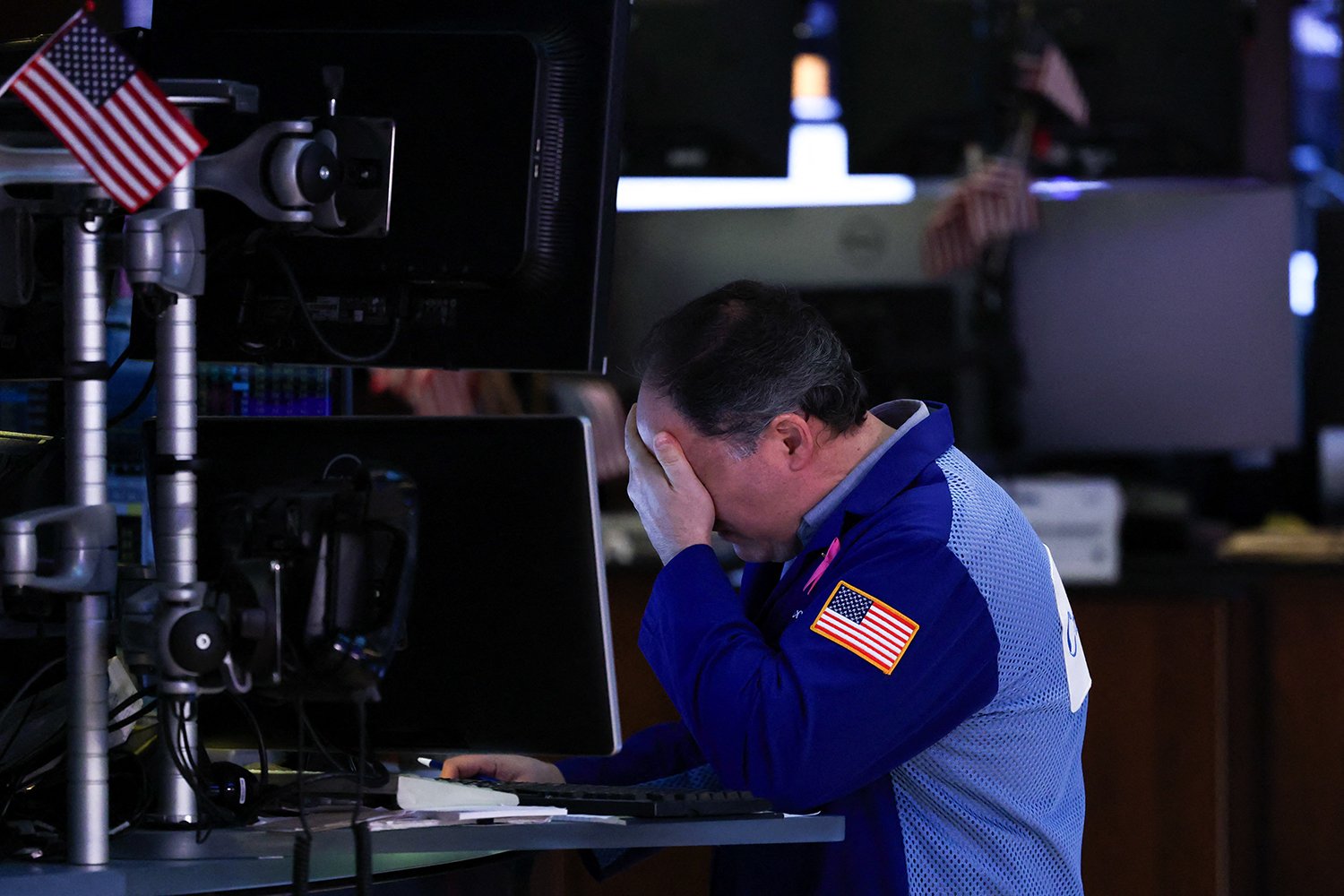 A trader with a U.S. flag patch on his sleeve puts his head in his hands as he sits in front of a computer monitor on the floor of the New York Stock Exchange.