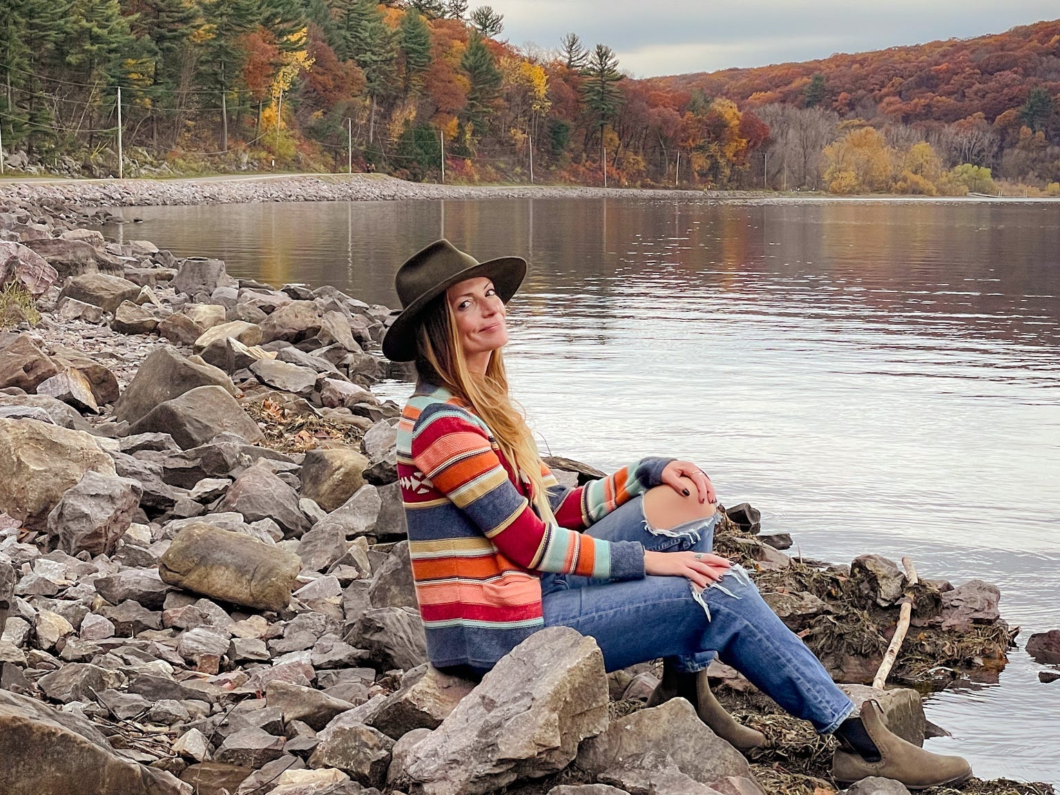Emily sits on rocks near a lake, with fall foliage in the background.