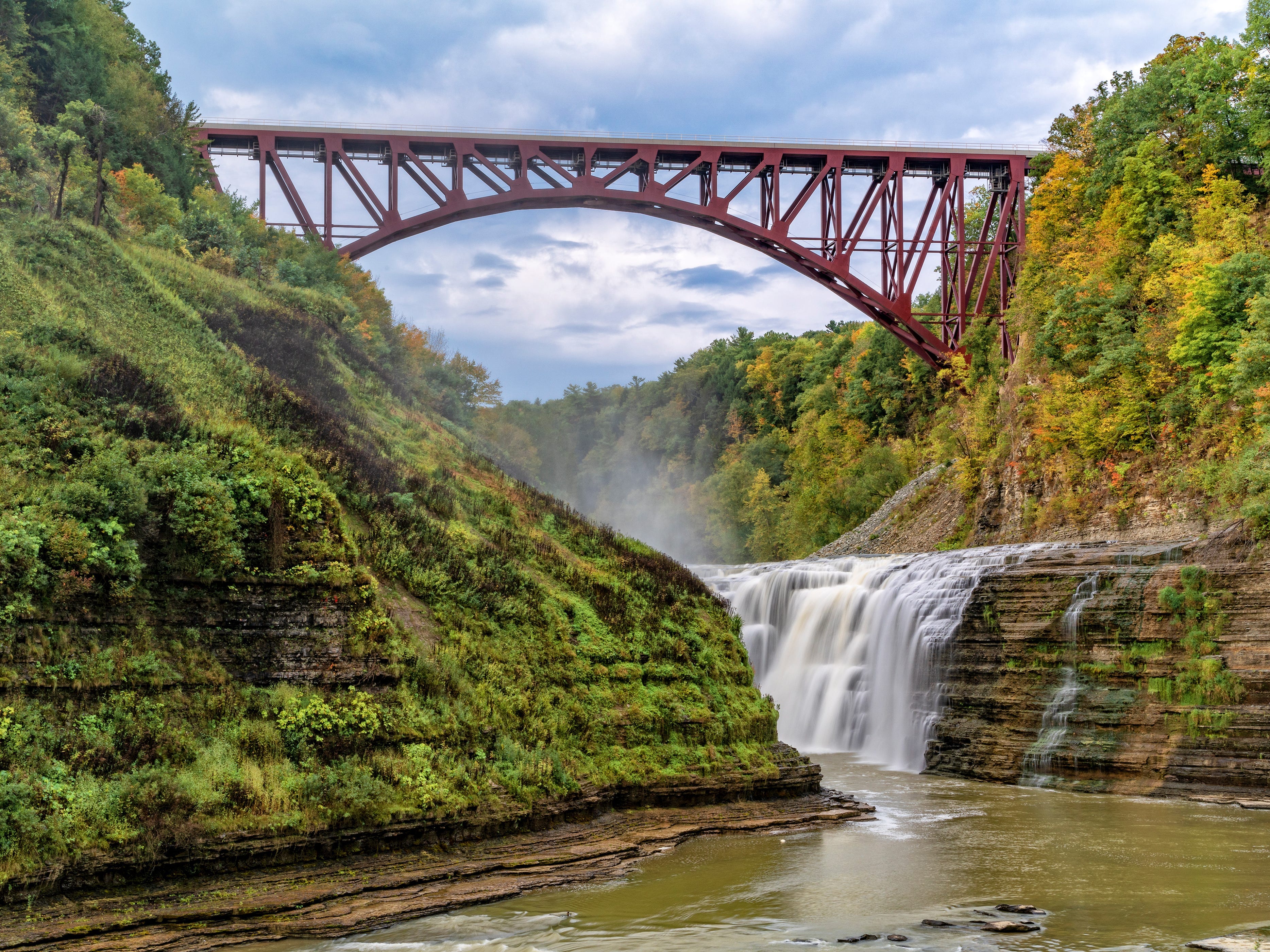 A curved bridge over trees and a waterfall.