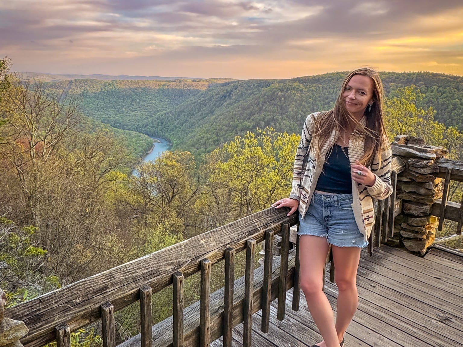 Emily stands at an overlook near a river and tree-covered mountains at sunset.