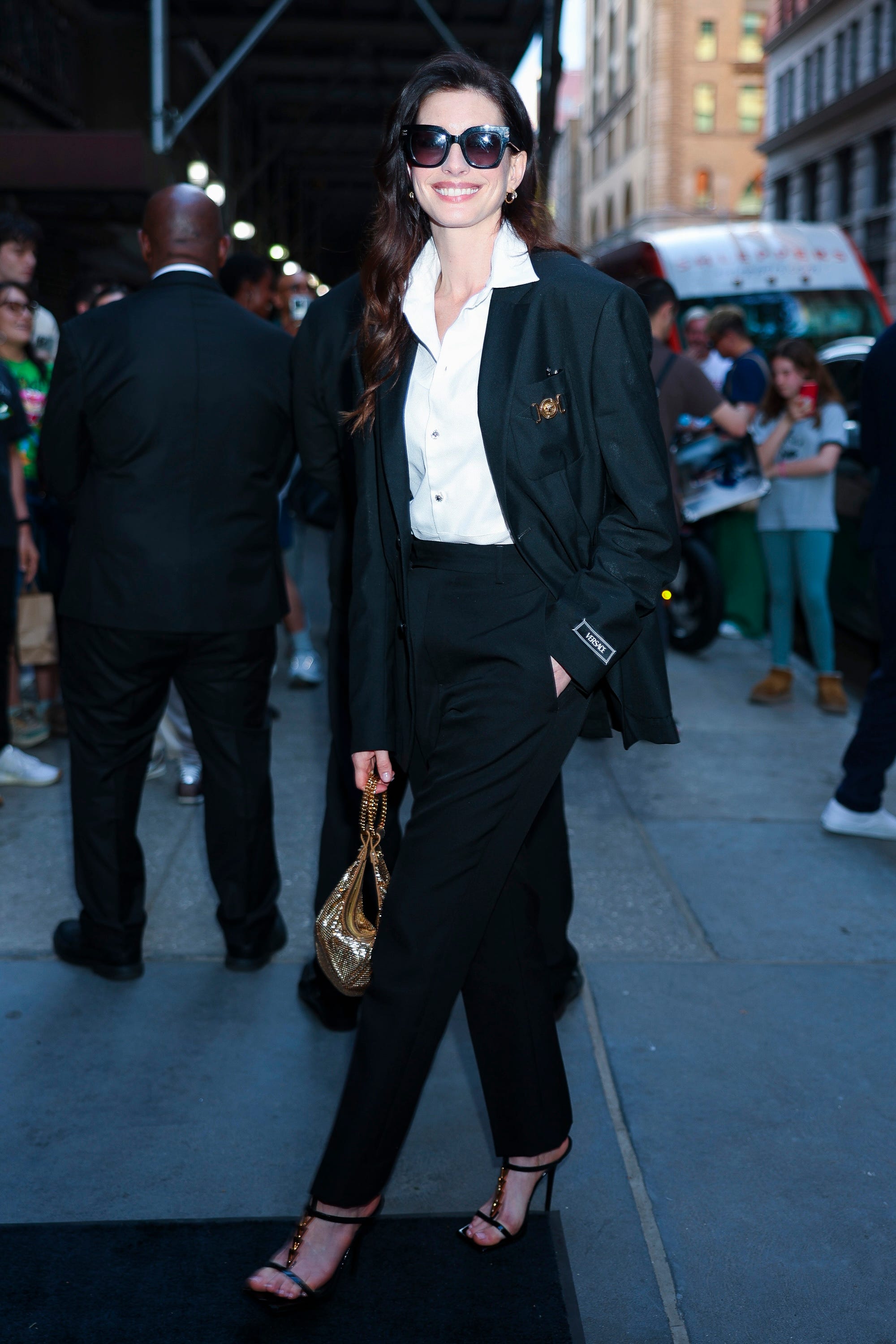 Anne Hathaway stands in a black suit and sunglasses.
