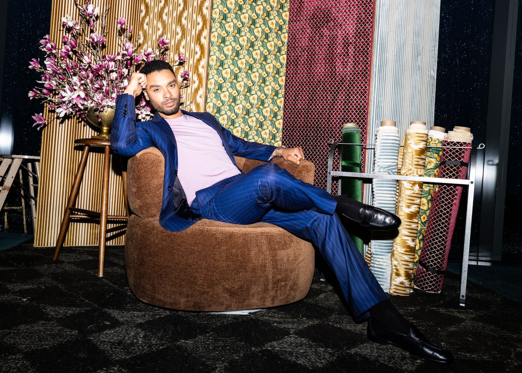 Regé-Jean Page sits in a blue suit on a brown chair.