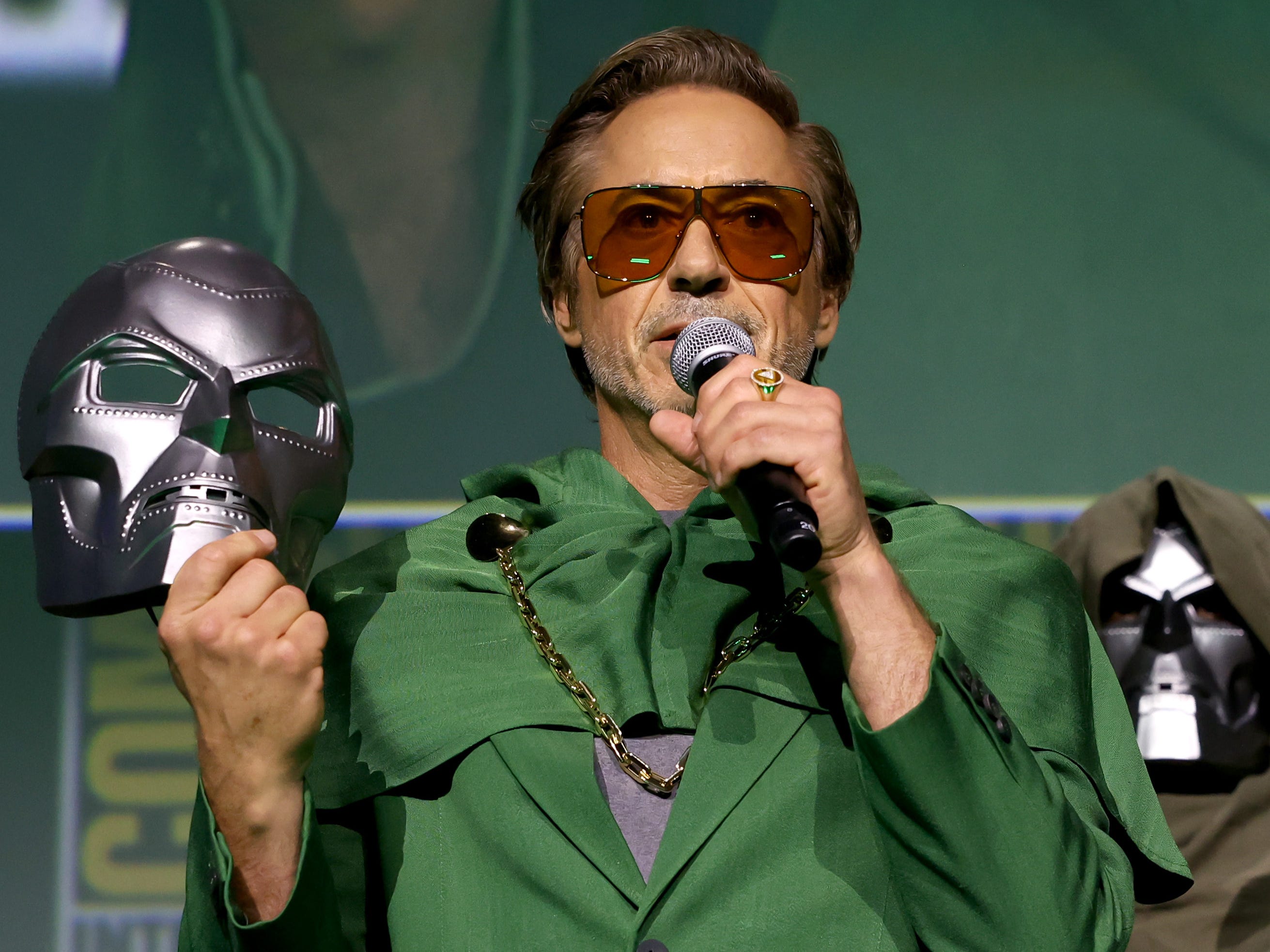 Robert Downey Jr. in a green suit holding a silver mask and a microphone.