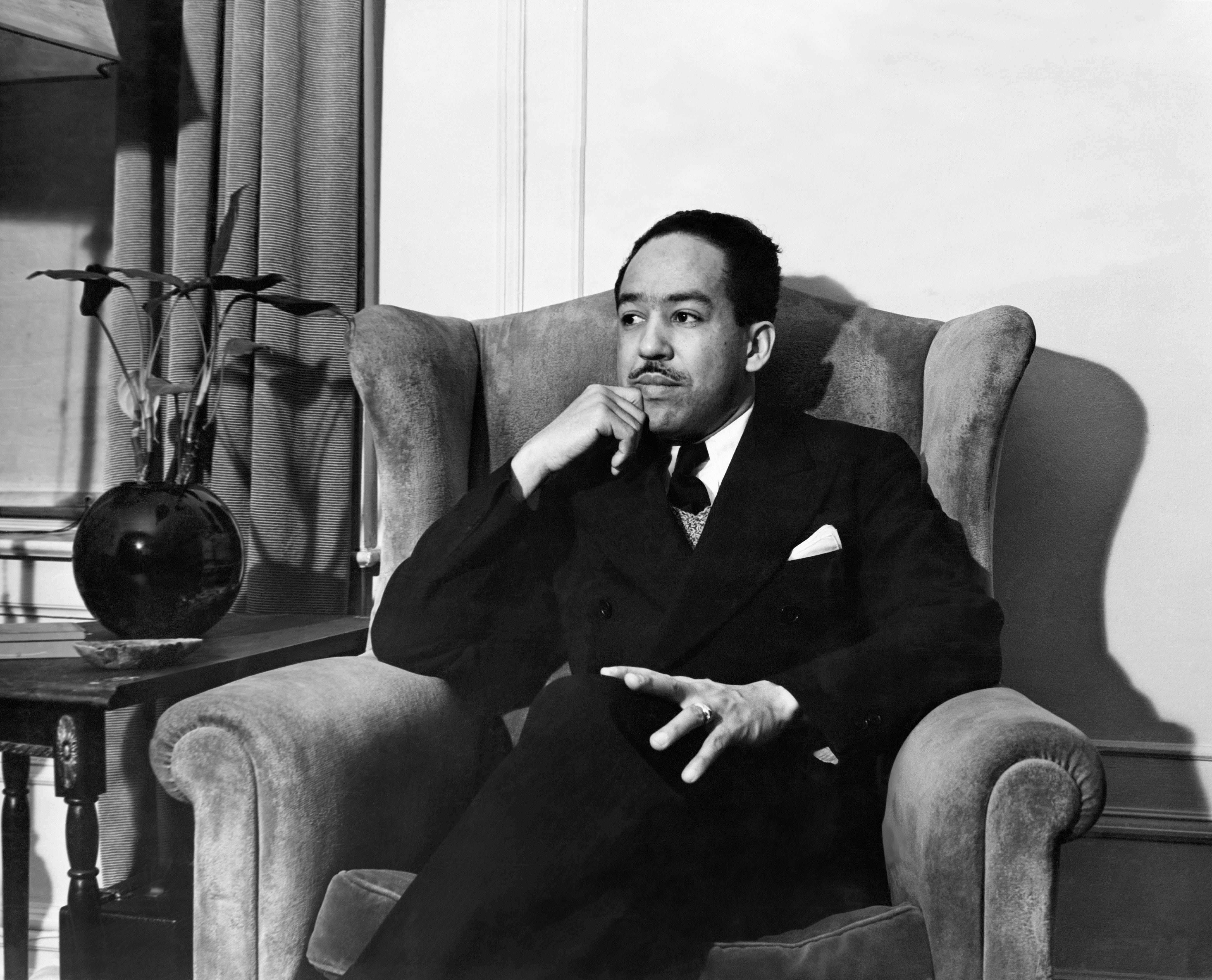 Langston Hughes sits in a chair while wearing a black suit and tie.