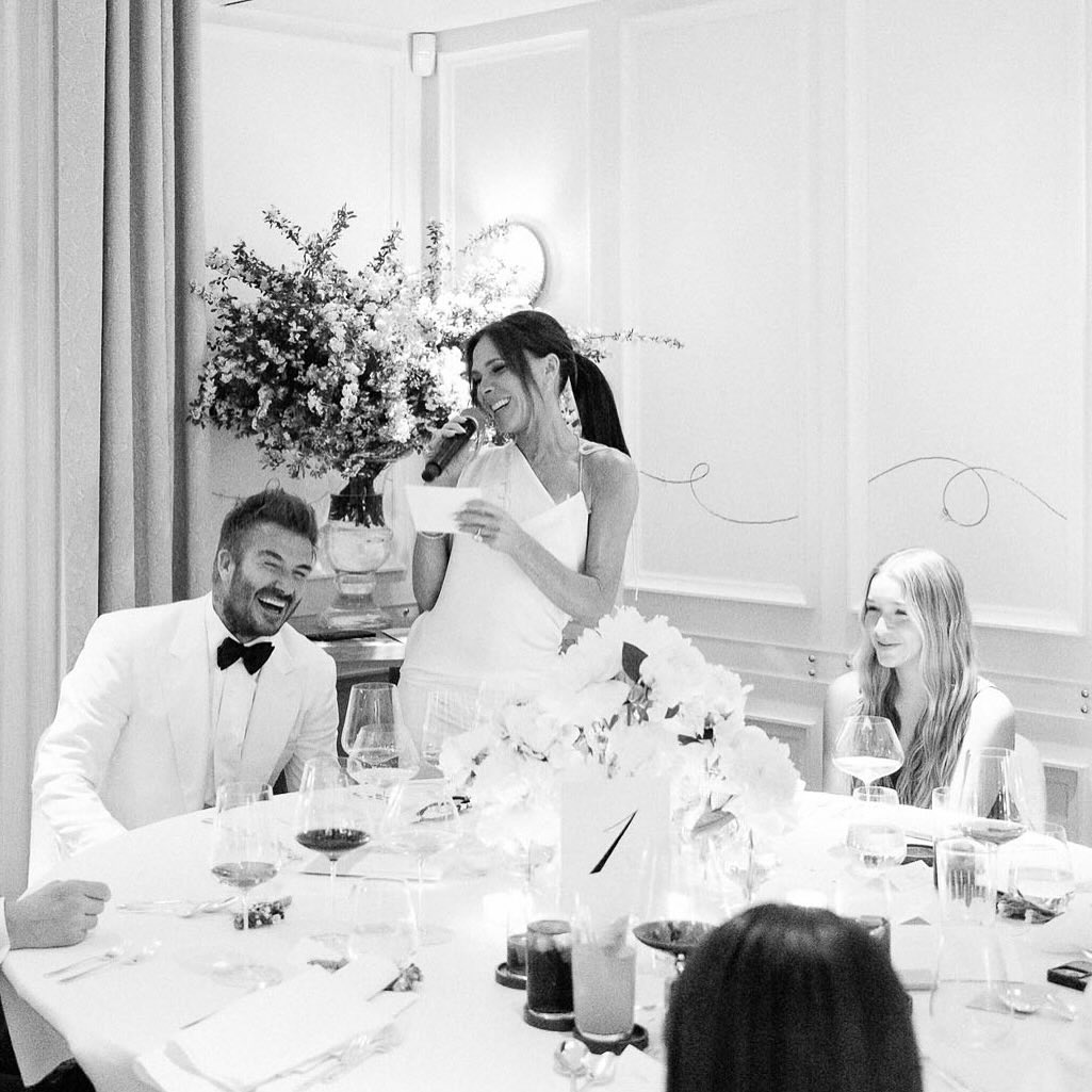 Black and white photo of David and Harper Beckham sitting at a table and Victoria Beckham standing, and all are laughing