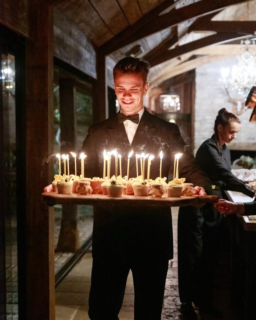 Romeo Beckham in a black tuxedo, white shirt and black bow tie, carrying a platter of cupcakes and lit candles.