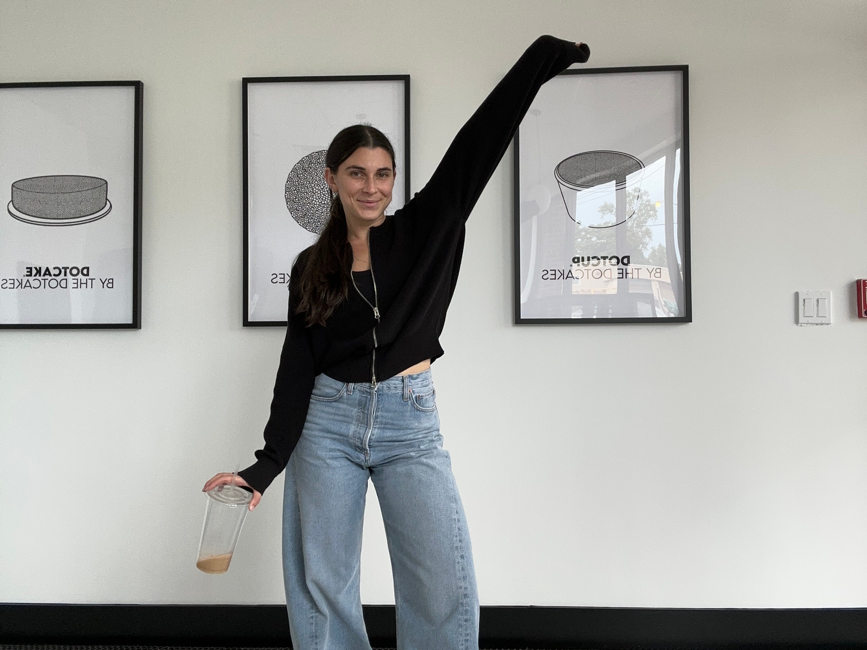 Alex Posner poses in front of black-and-white illustrations of Dotcakes in picture frames on the wall behind her, with one hand in the air and the other holding a drink down by her right thigh.